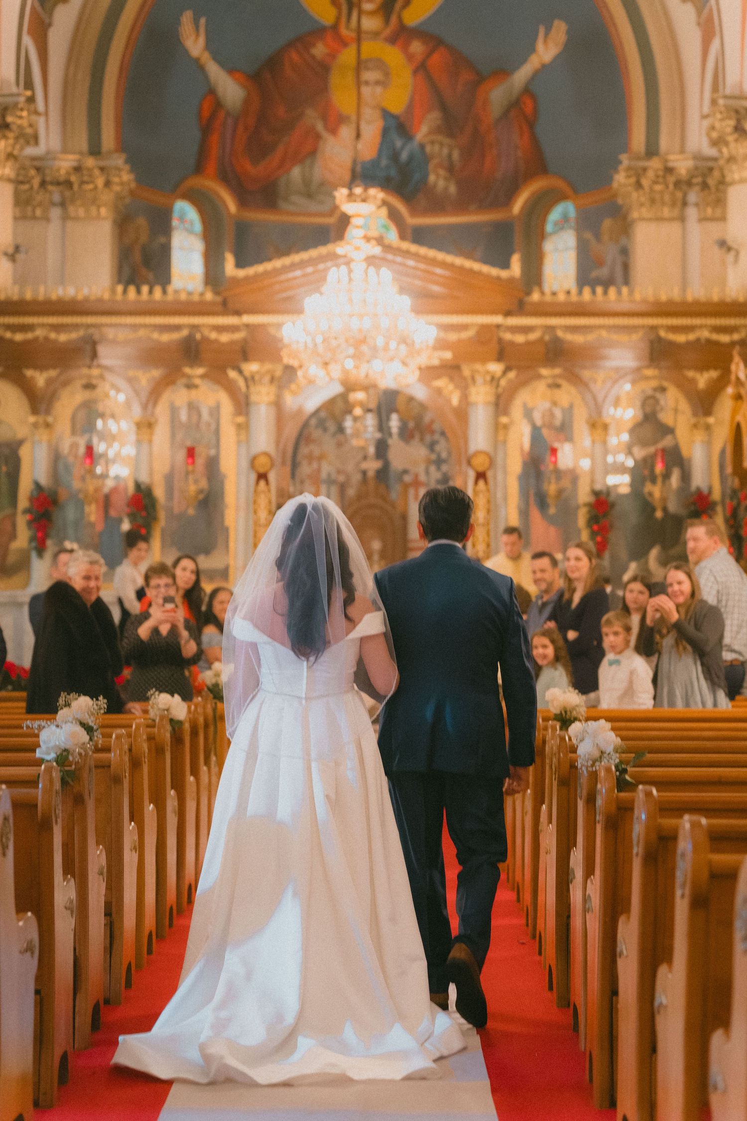 Bride and groom walk down red carpet aisle of ornate Orthodox church decorated with religious icons and Christmas decor.