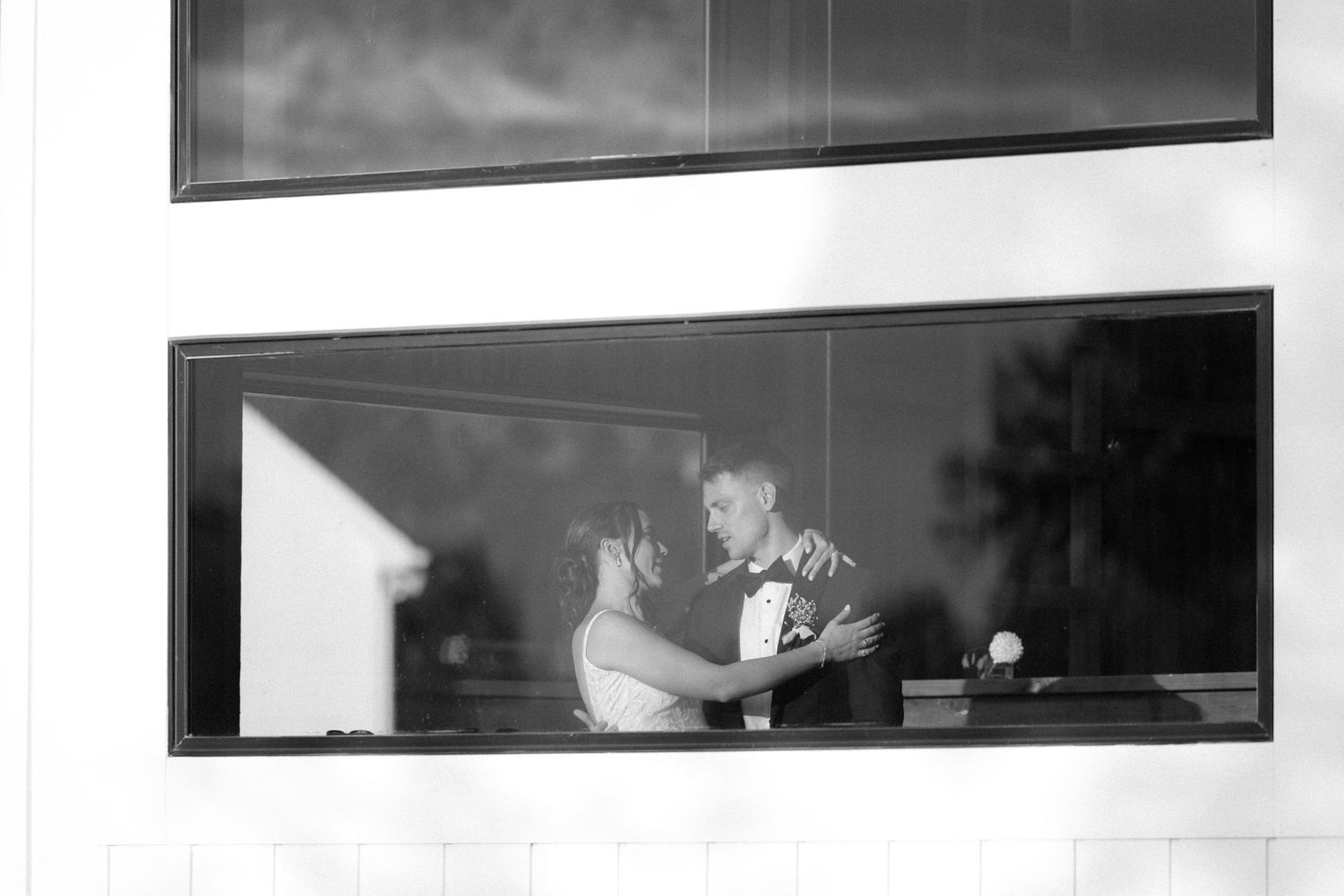 Black and white photo of bride and groom embracing romantically reflected in large modern window.