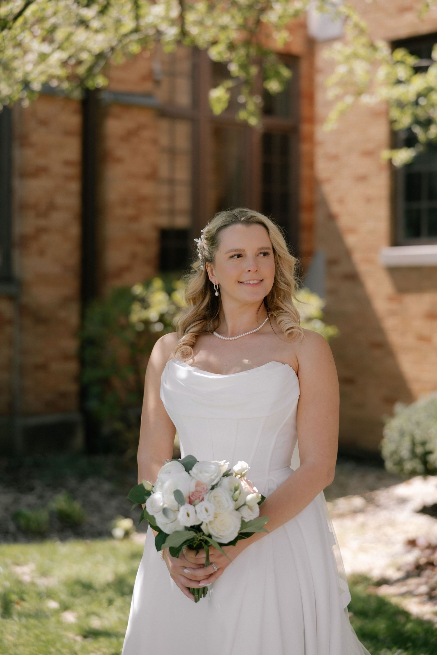 Bride in strapless white gown holding white floral bouquet, smiling outdoors near brick building on sunny day.