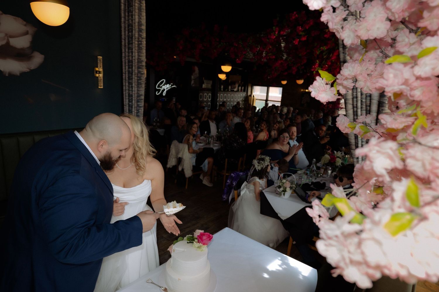 Bride and groom cutting their white wedding cake surrounded by pink cherry blossom decorations and wedding guests.