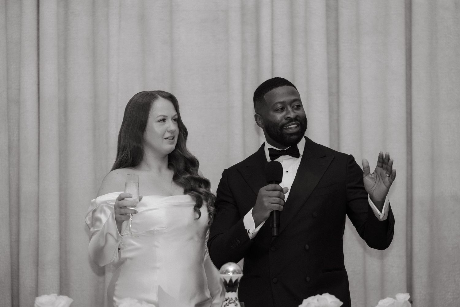 Black and white photo of a bride and groom speaking at their wedding reception, holding microphone and champagne glass.