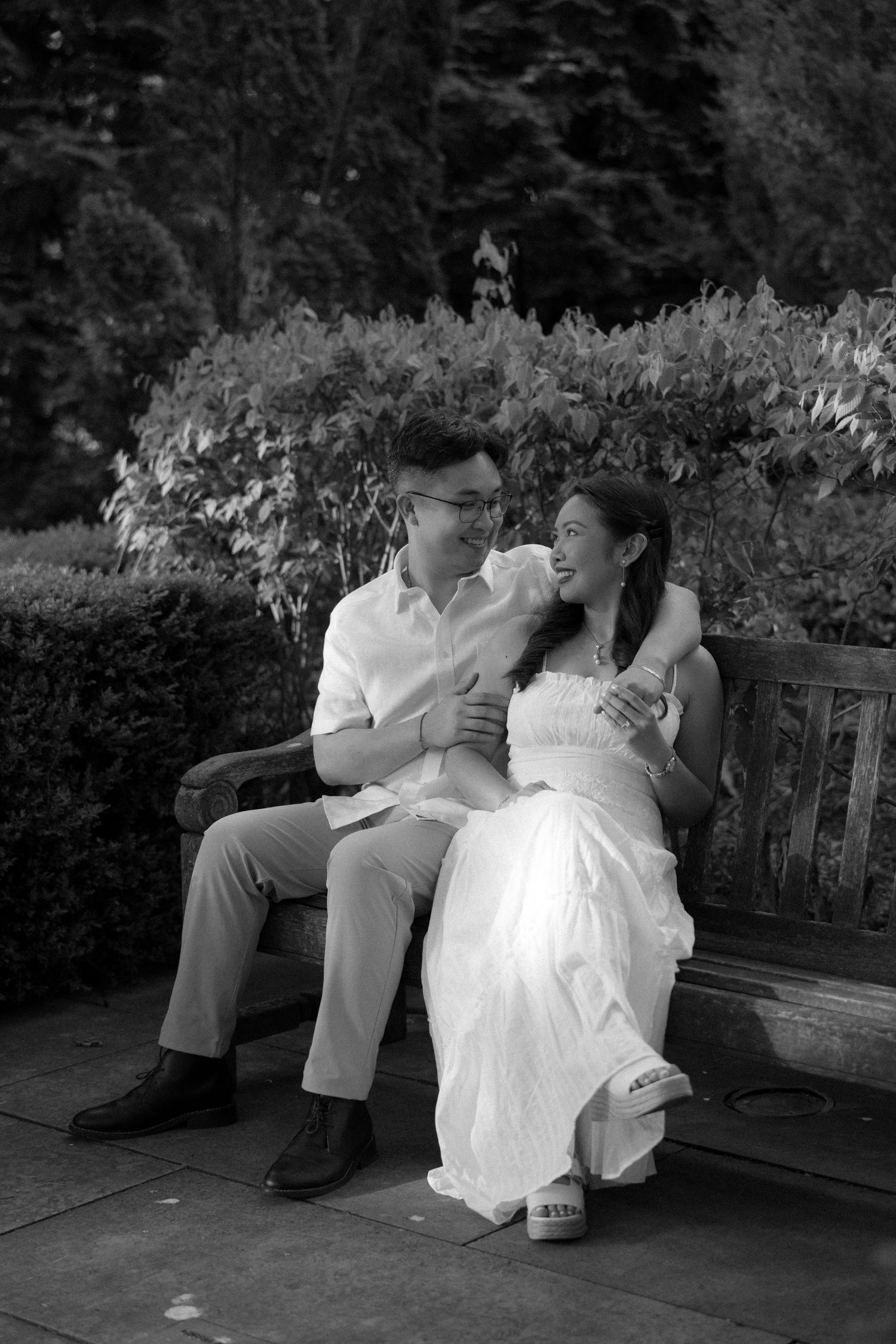 Black and white photo of a couple in wedding attire sitting on a bench in a garden, smiling at each other.