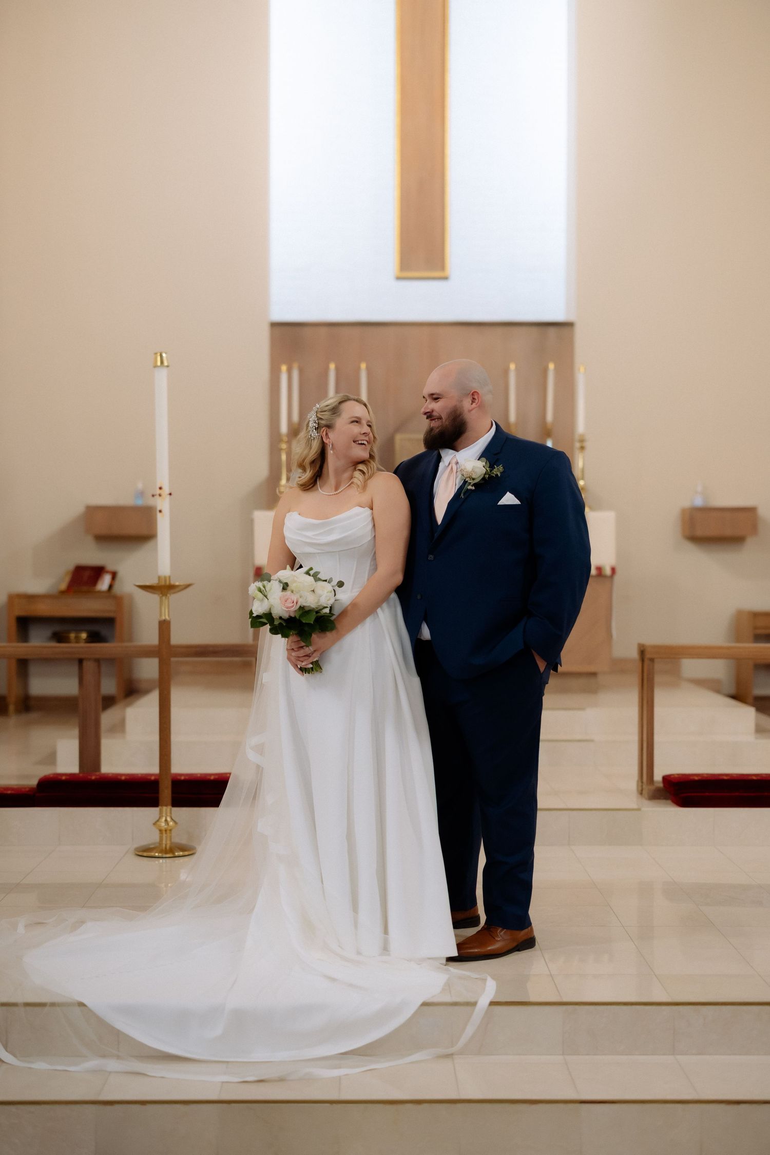 Bride and groom smiling together at church altar, bride in white gown holding bouquet, groom in navy suit.
