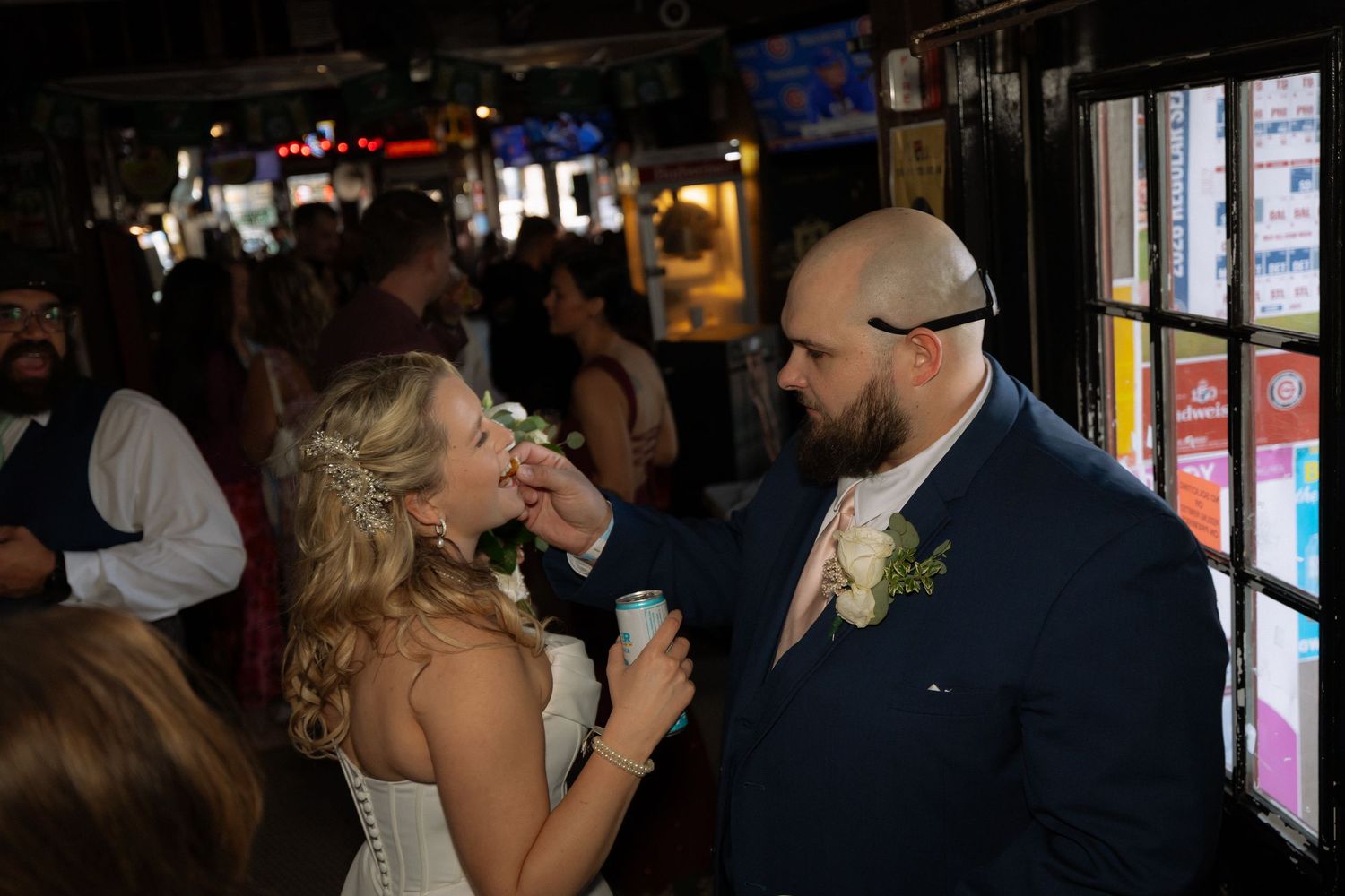Groom feeding bride wedding cake at reception venue, bride in white gown with floral hairpiece, groom in navy suit.