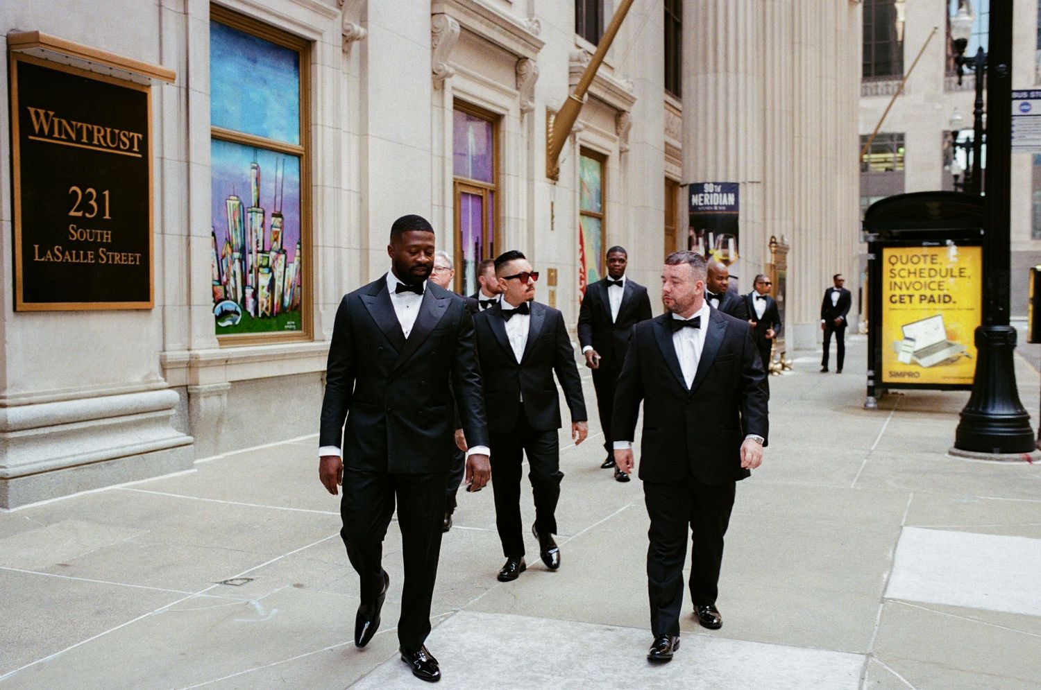 Four groomsmen in black tuxedos and bow ties walk confidently outside Wintrust building at 231 N LaSalle Street Chicago.