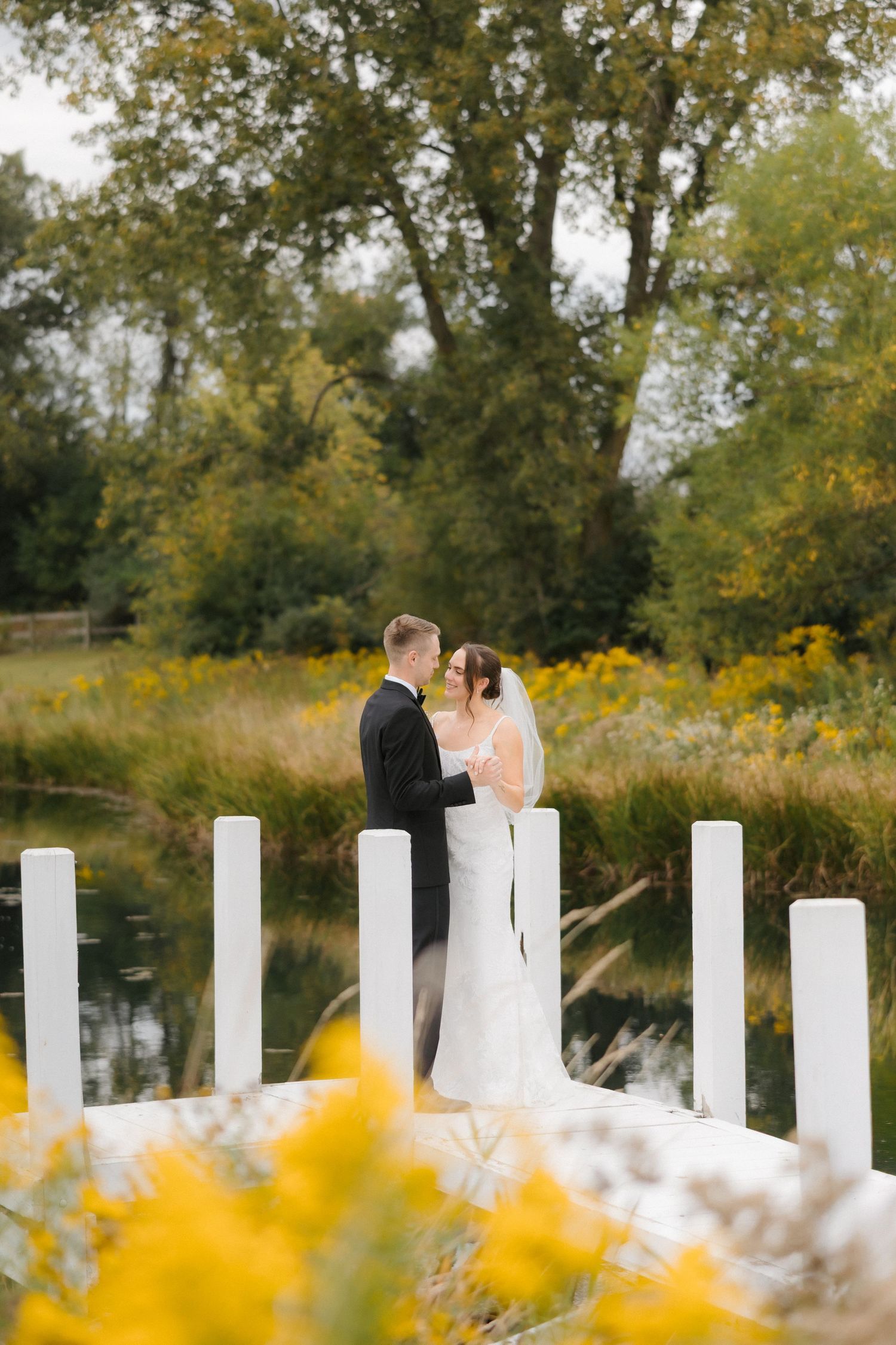 Bride and groom sharing an intimate moment on a white dock framed by yellow wildflowers and a pond.