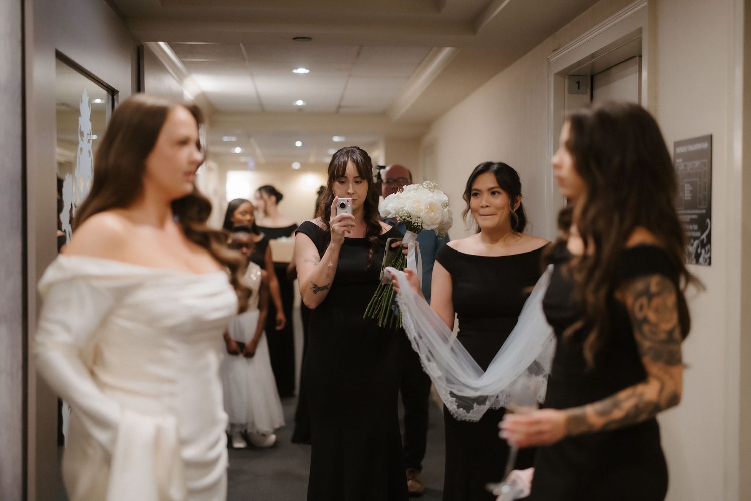 Bridesmaids in black dresses gather in a hallway as bride in white gown prepares, one holding white roses bouquet.