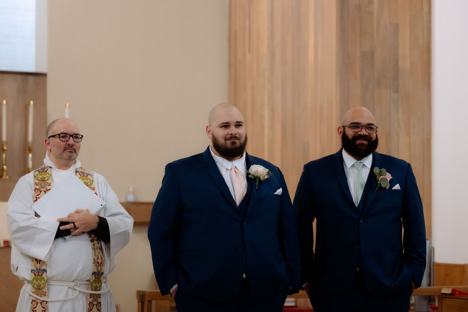 Groom in navy suit stands at church altar with best man and officiant in white robes during wedding ceremony.