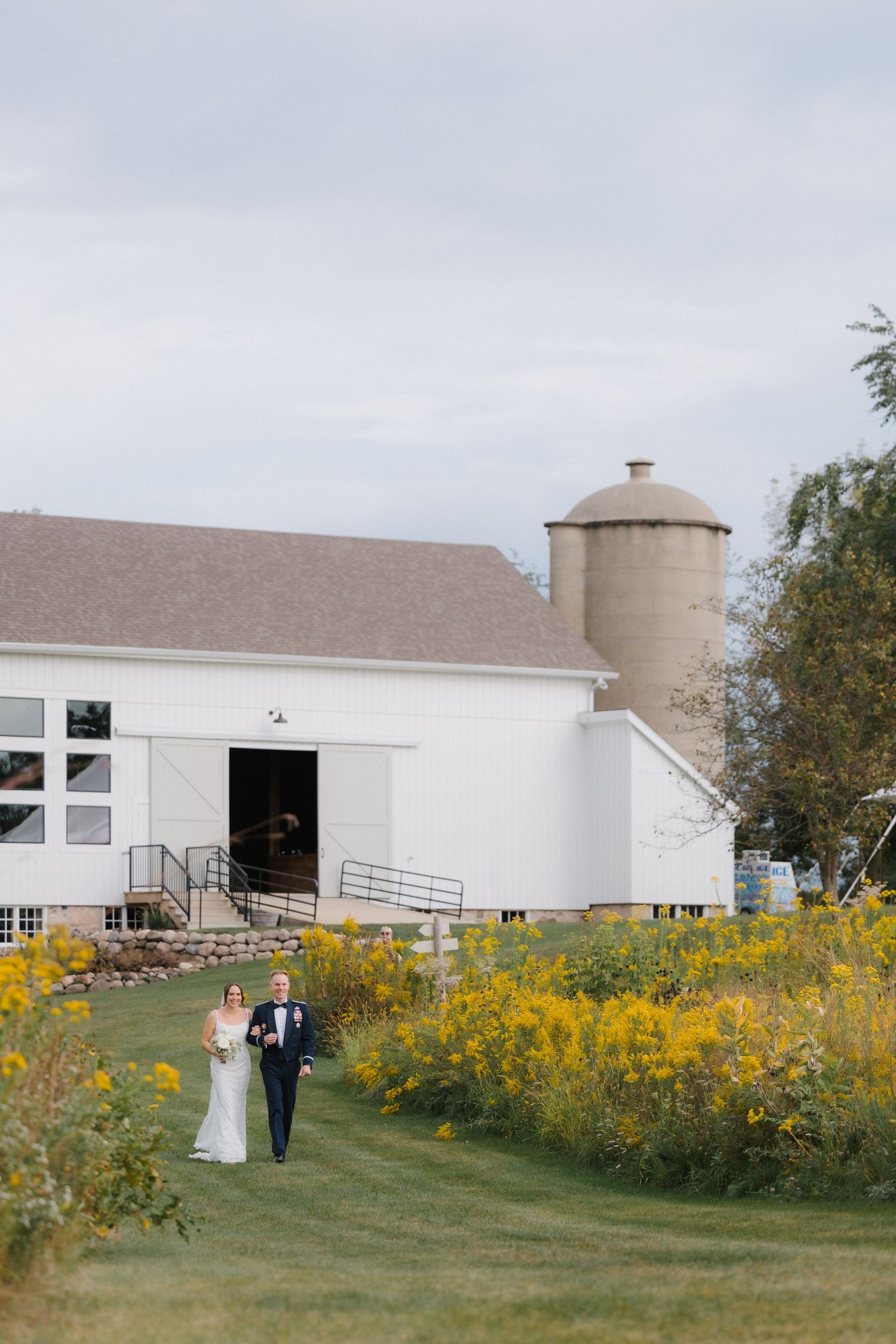 Bride and groom walk through golden wildflower meadow beside a white barn venue with silo on their wedding day.
