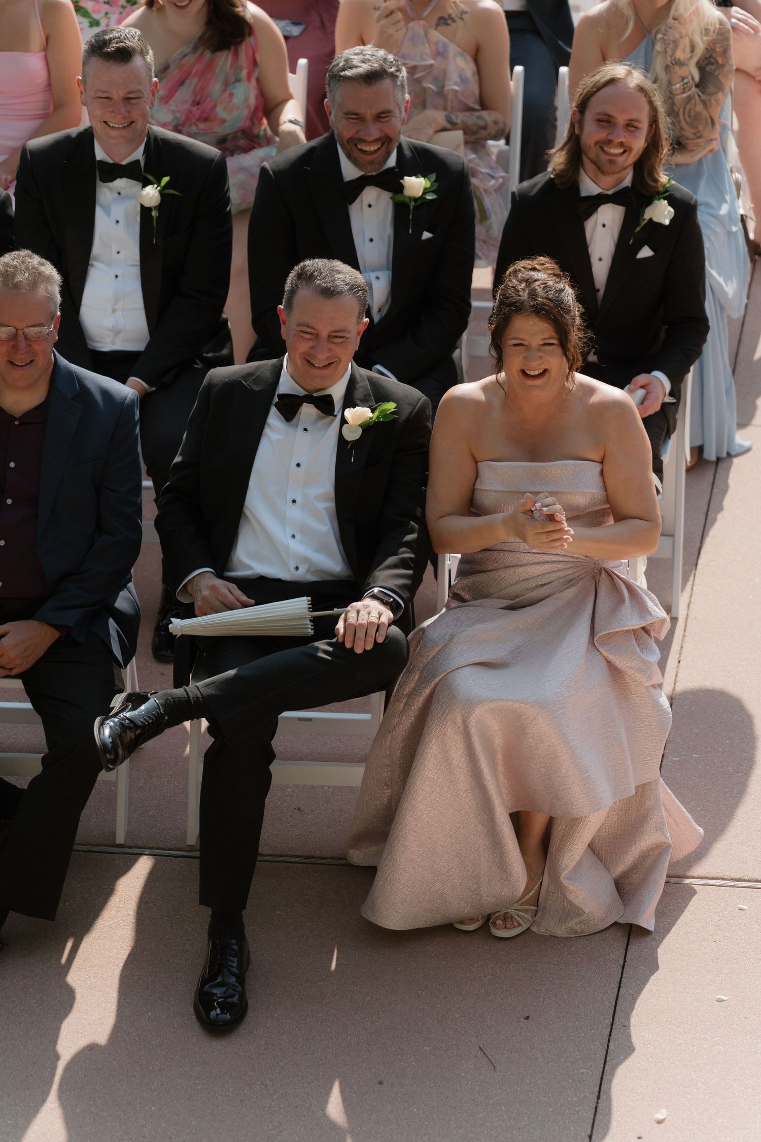 Wedding guests laugh joyfully during an outdoor ceremony, with groomsmen in tuxedos seated in white chairs on a sunny day.