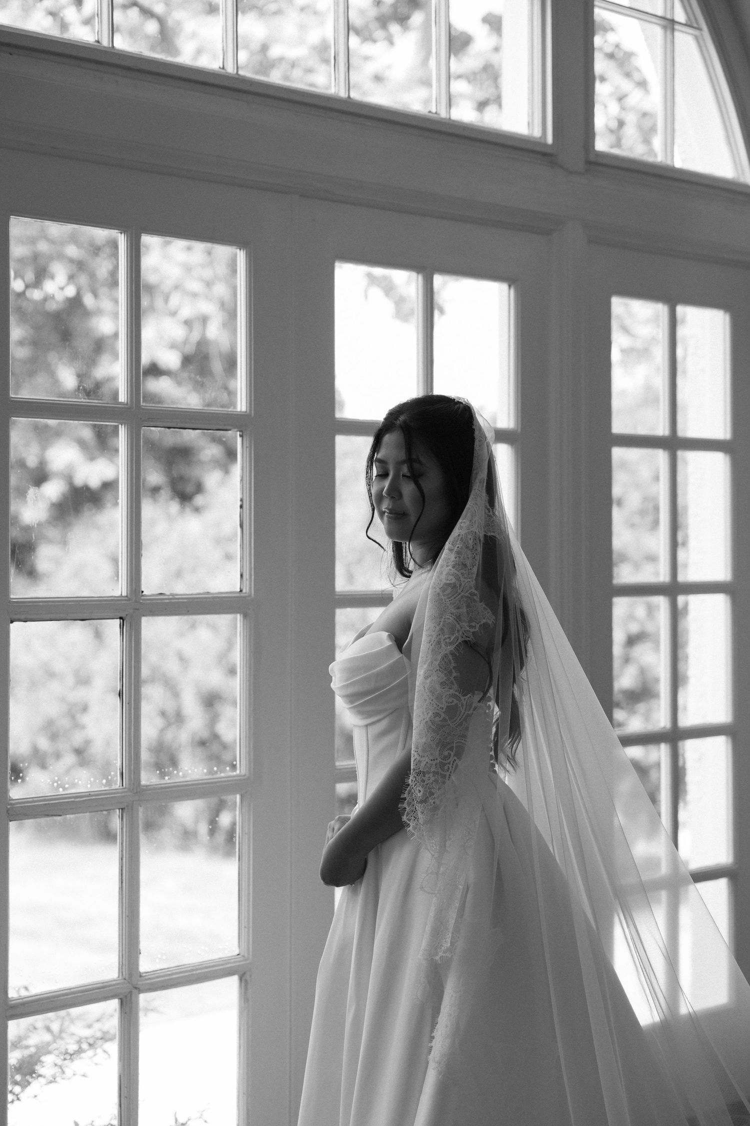 Elegant bride in wedding gown and lace veil standing by large French windows in black and white photo.