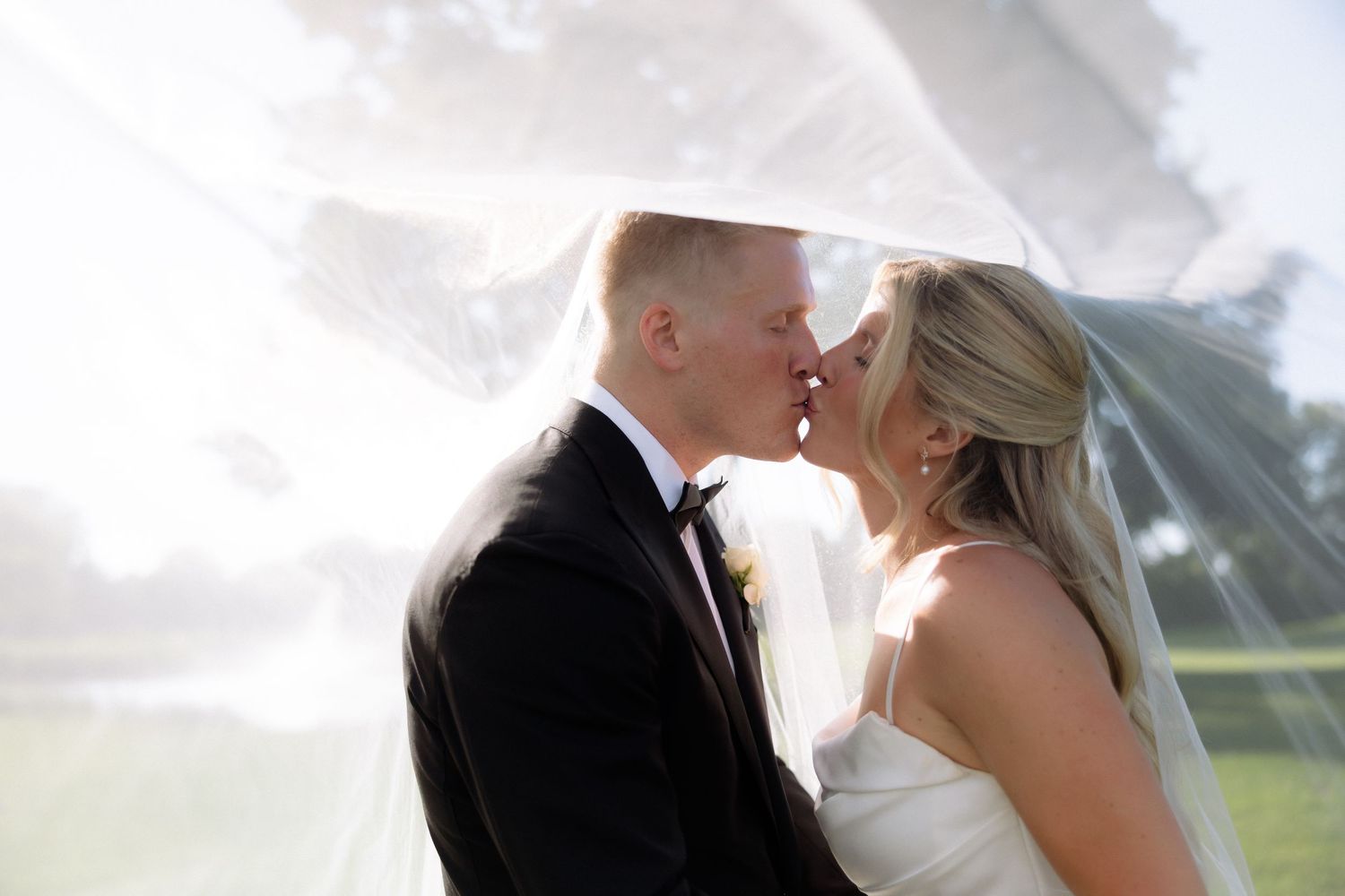 Bride and groom kissing under flowing veil with sunlight streaming through at outdoor wedding.