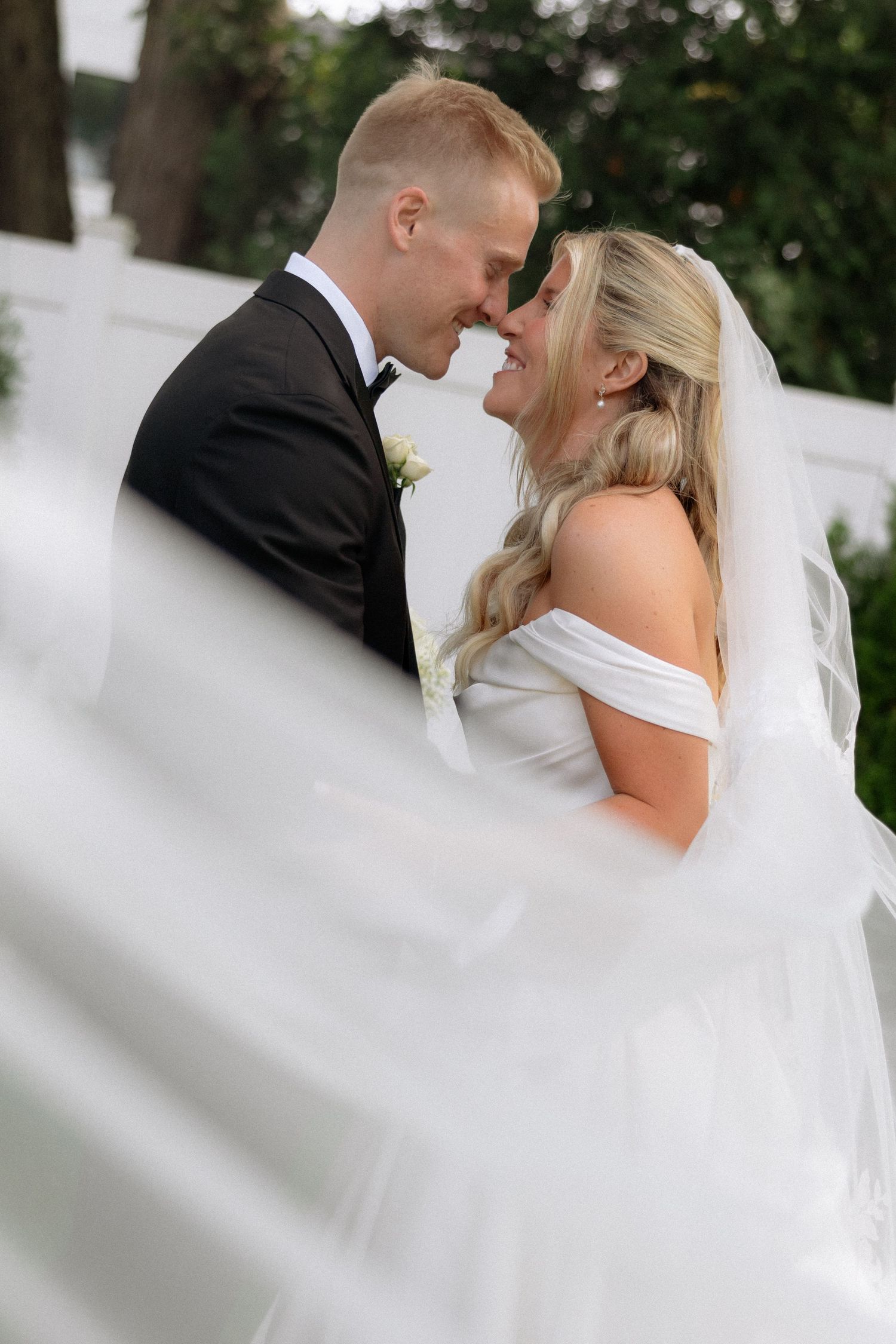 Bride and groom share romantic nose-to-nose moment as white veil billows around them outdoors.
