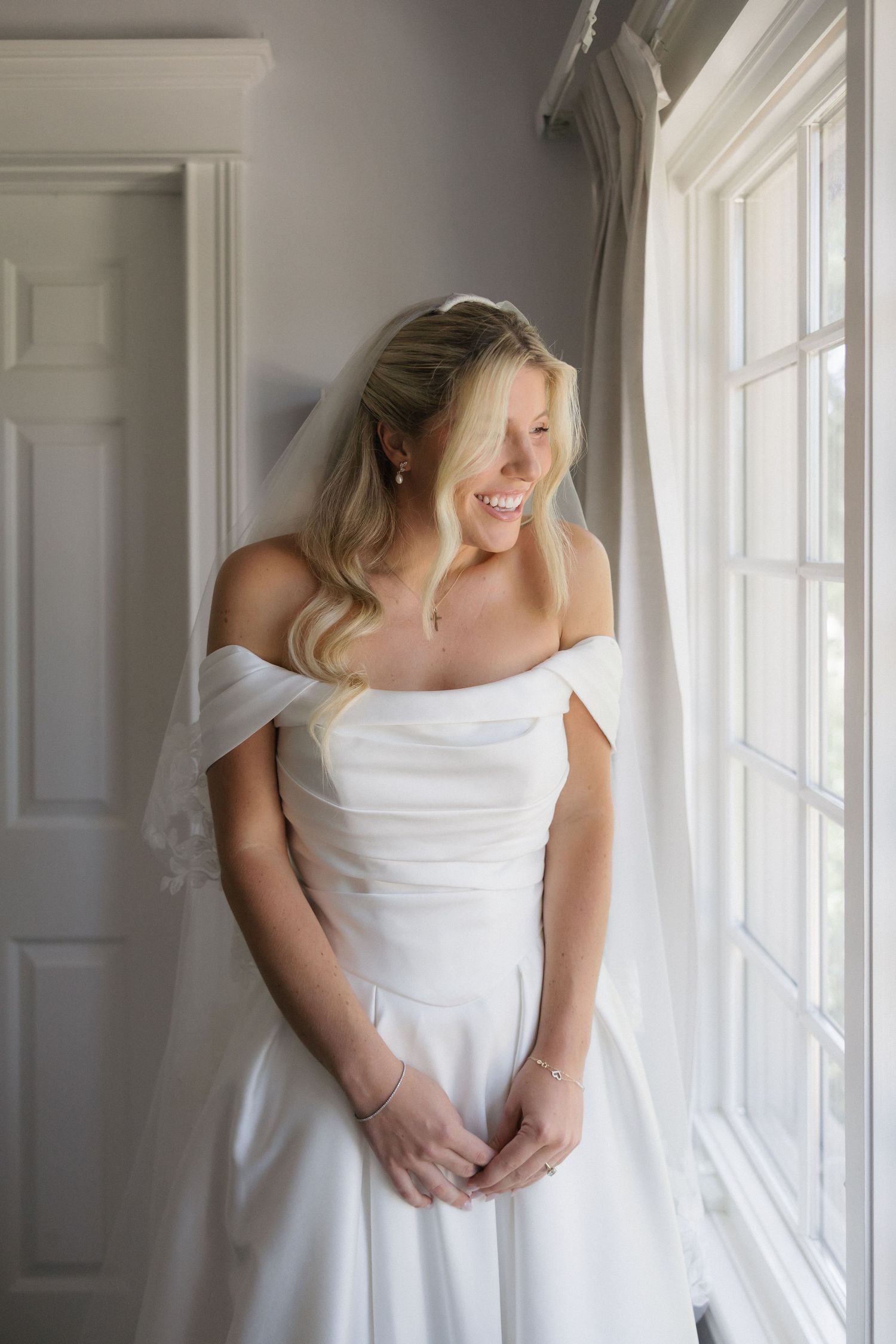 Smiling bride in off-shoulder white wedding gown and veil standing by a bright window, hands clasped.