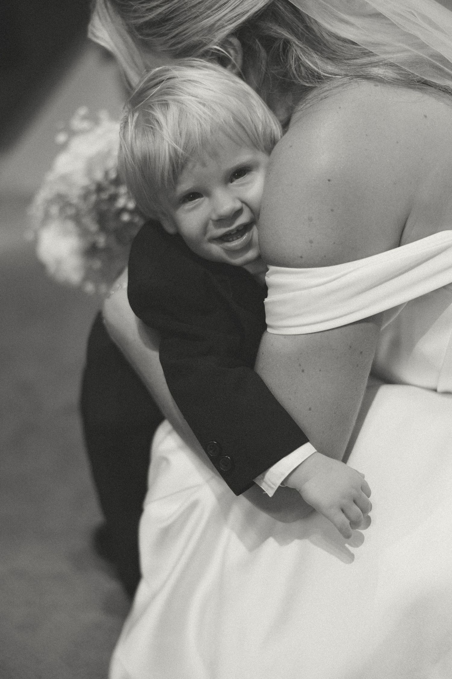 Smiling toddler boy in tuxedo hugging bride in white wedding dress, black and white photo.