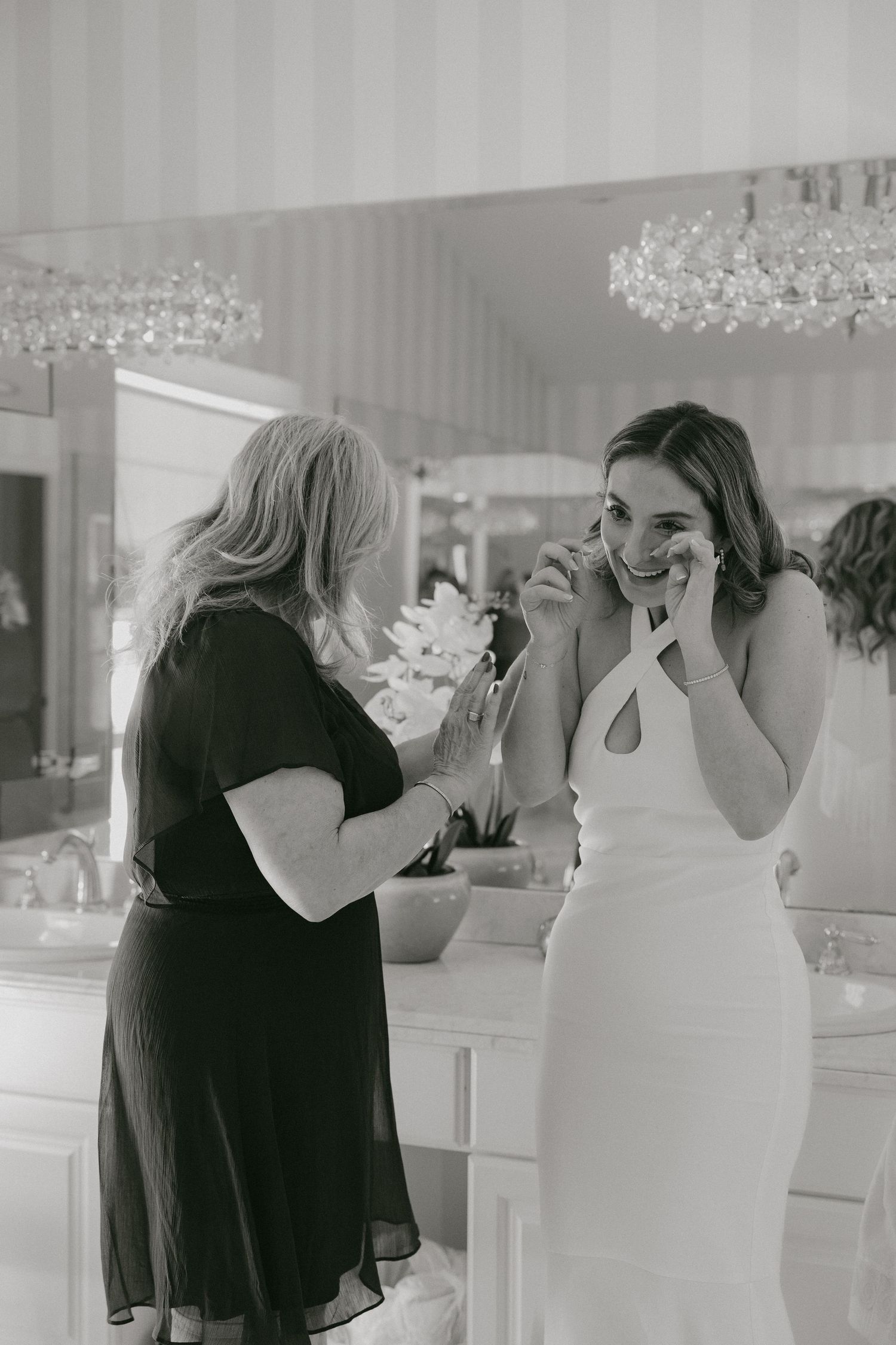 Emotional black and white photo of bride wiping tears while woman assists her in a mirrored bathroom.