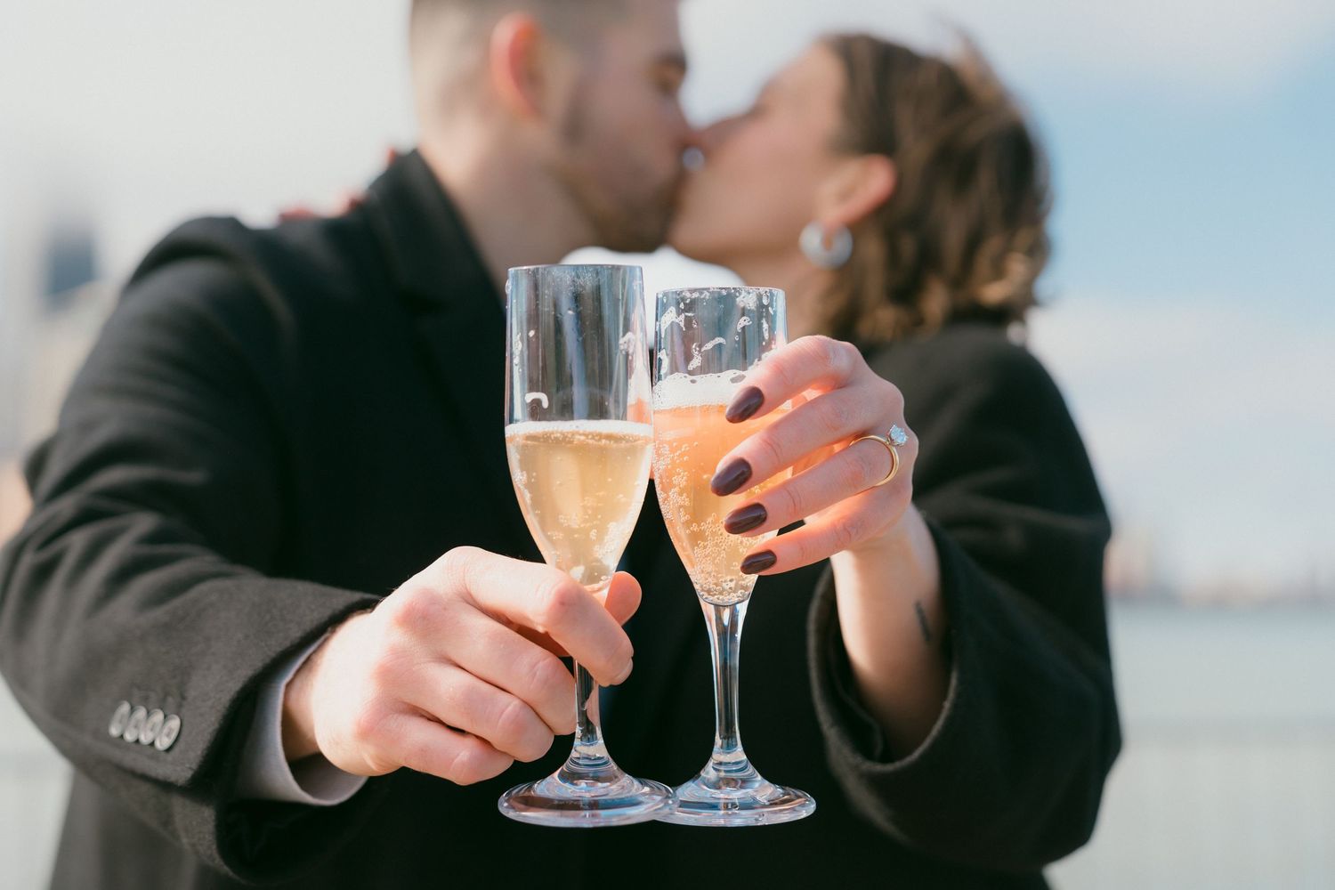 Engaged couple kissing behind two clinking champagne flutes with a city skyline in the background.