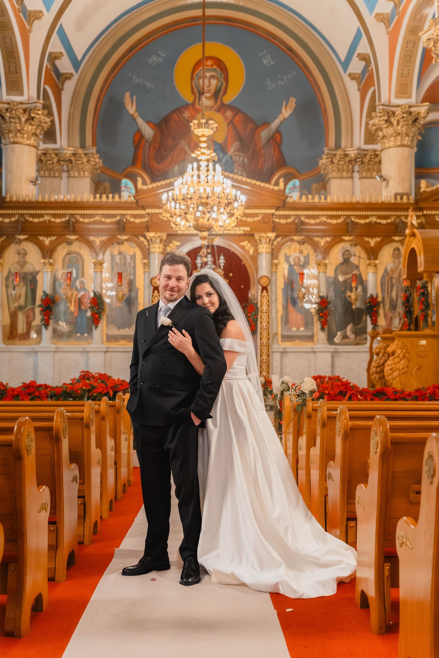 Bride and groom pose together on red carpet aisle of stunning Orthodox church with golden iconostasis and murals.