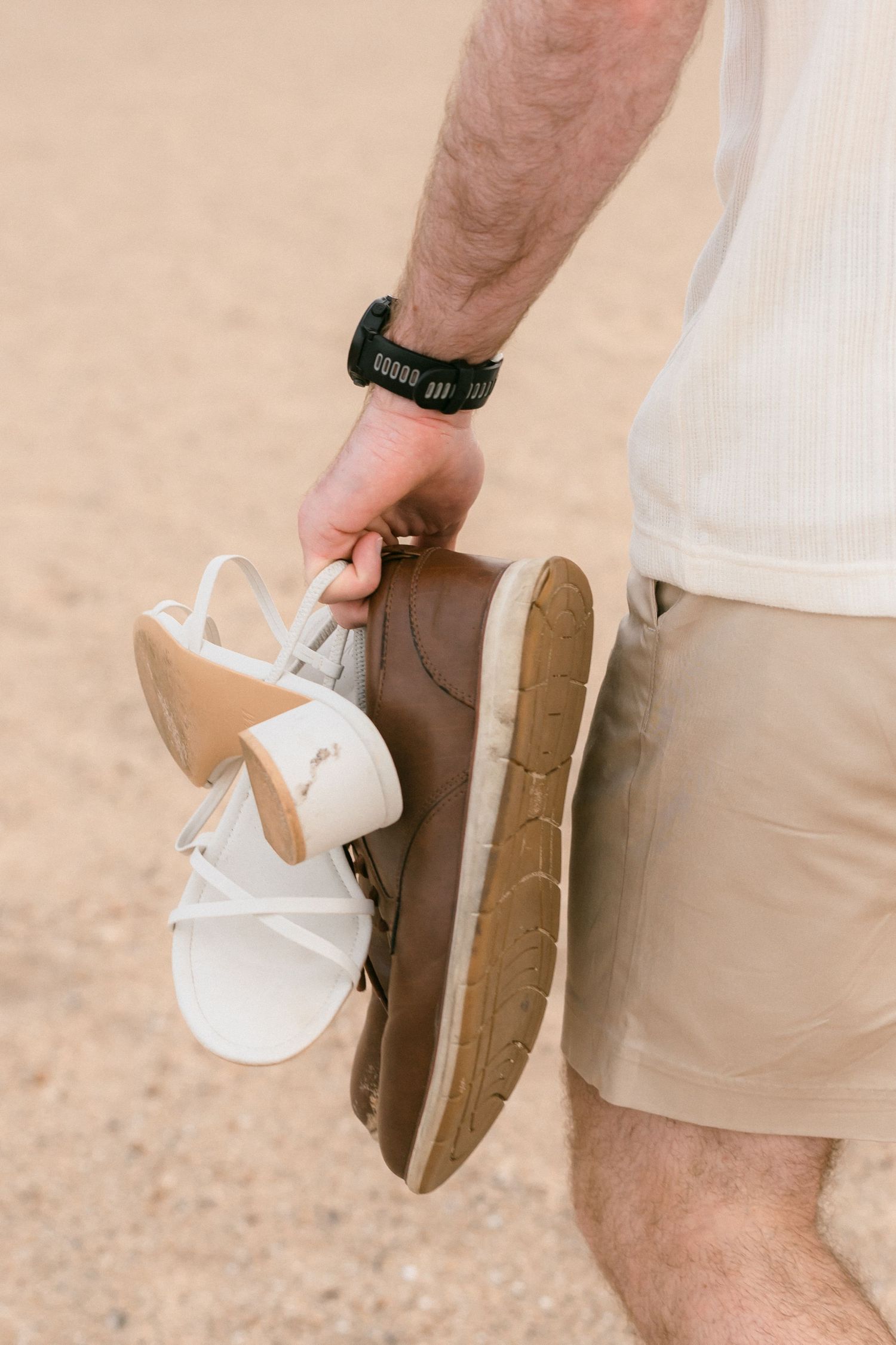 Close-up of a man's hand holding white sandals and brown shoes while walking on sandy beach.