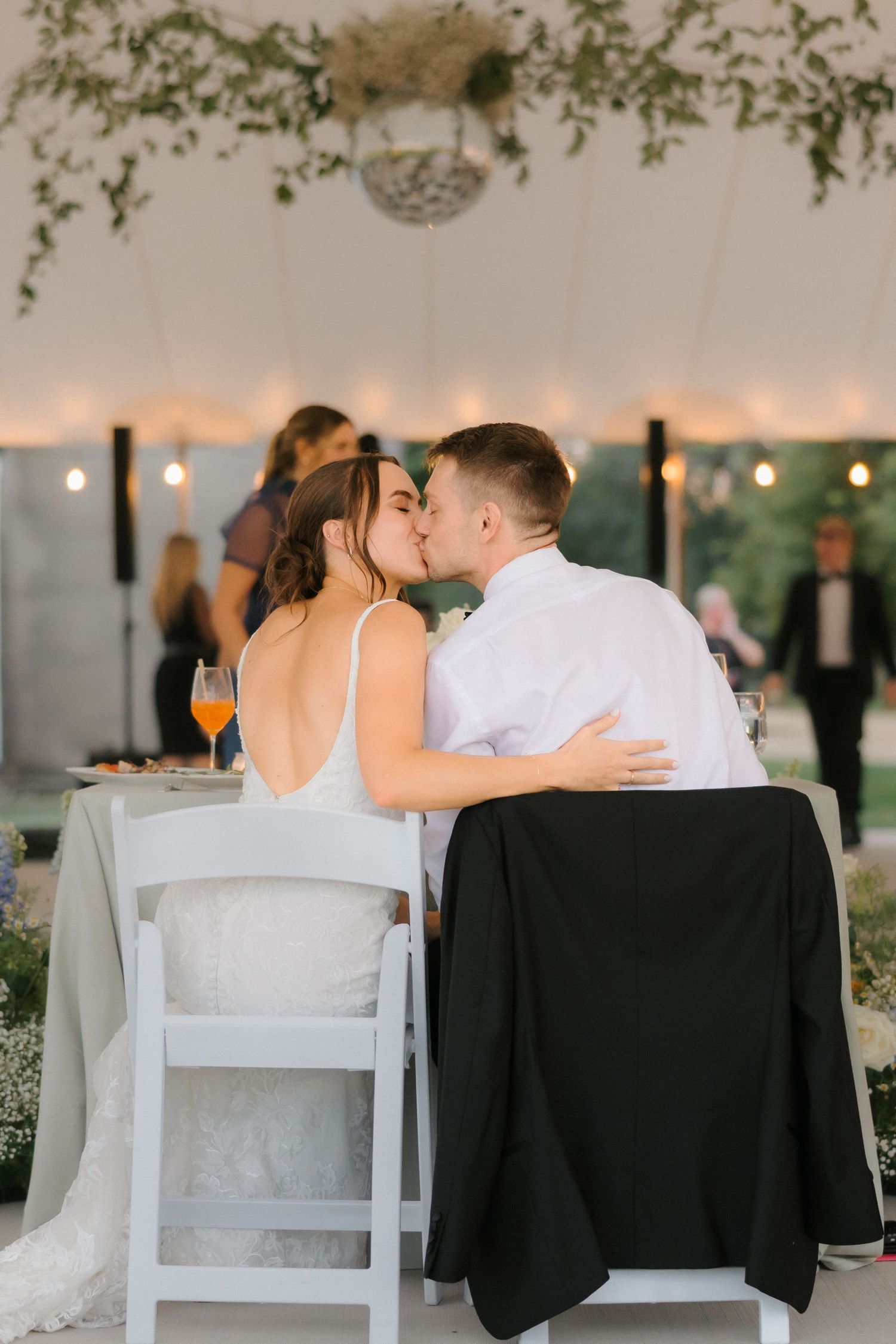 Bride and groom share a romantic kiss at their wedding reception table under string lights and greenery.