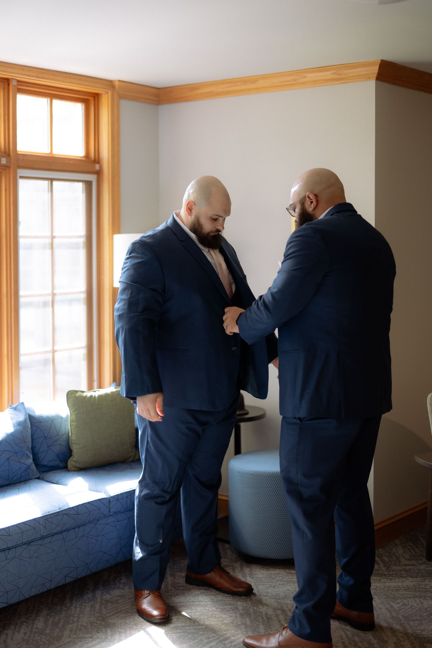 Two men in navy suits help each other get dressed in a bright room before a formal wedding ceremony.