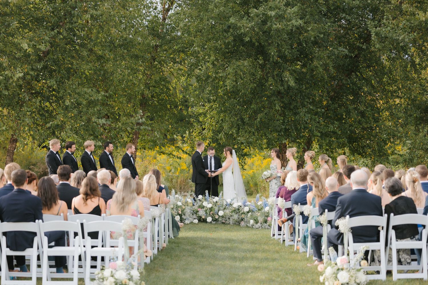 Wide view of elegant outdoor wedding ceremony with white chairs, floral arrangements lining the aisle, and lush green trees.