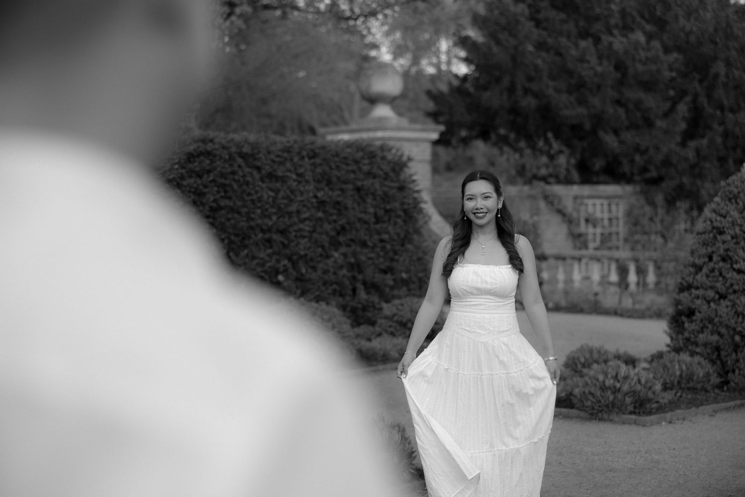 Black and white photo of a smiling bride in a strapless wedding gown walking through a garden toward the groom.
