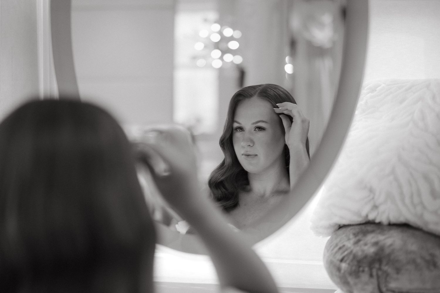 Black and white photo of a woman adjusting her hair while looking at her reflection in a round mirror.
