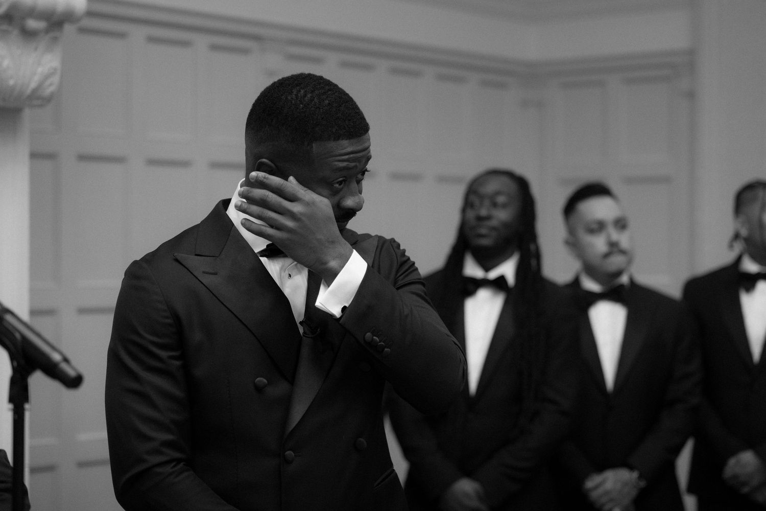 Emotional groom in black tuxedo wiping tears at wedding ceremony with groomsmen standing behind him.