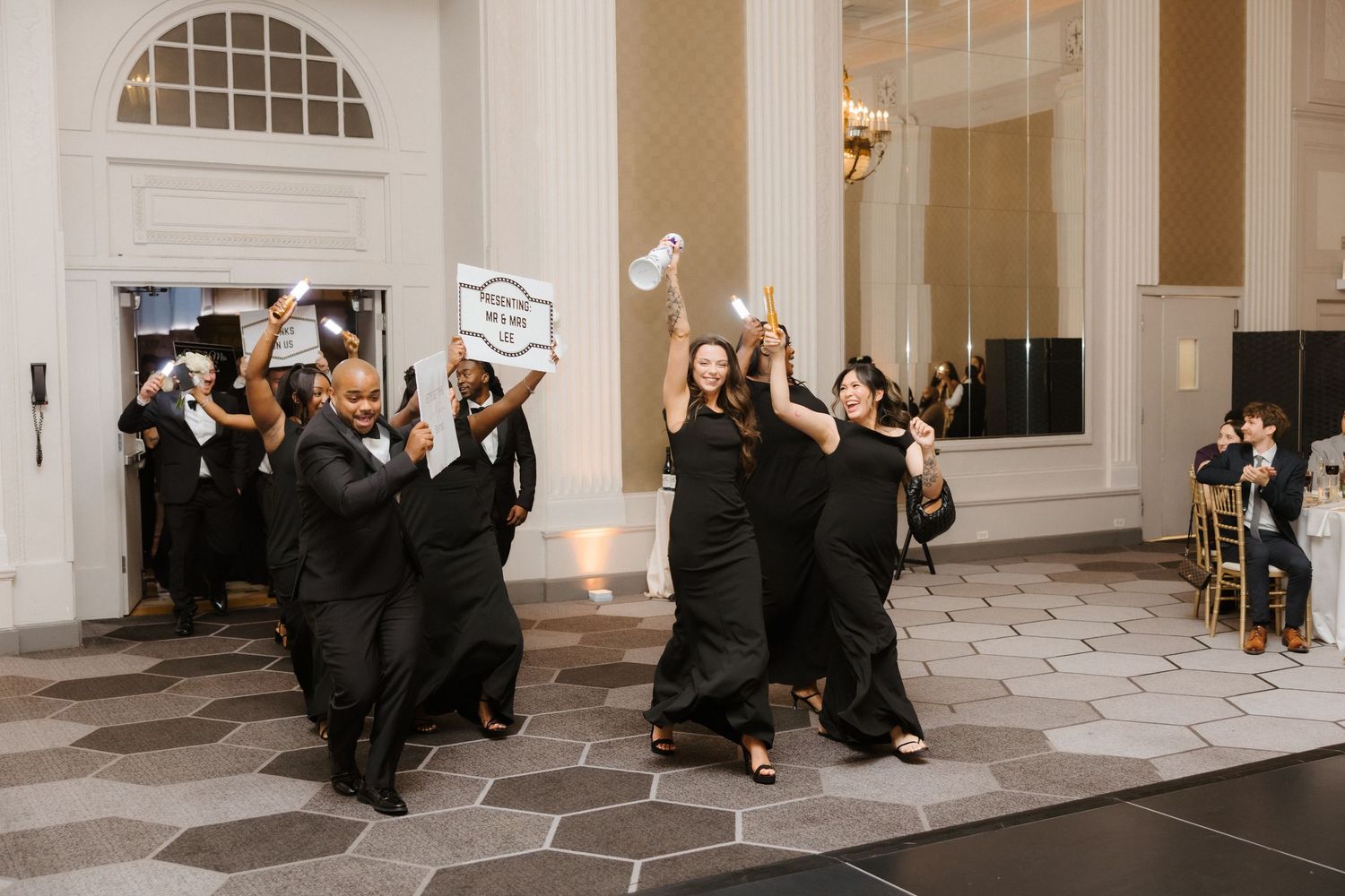 Bridesmaids in black gowns celebrate joyfully on a dance floor at an elegant wedding reception venue.