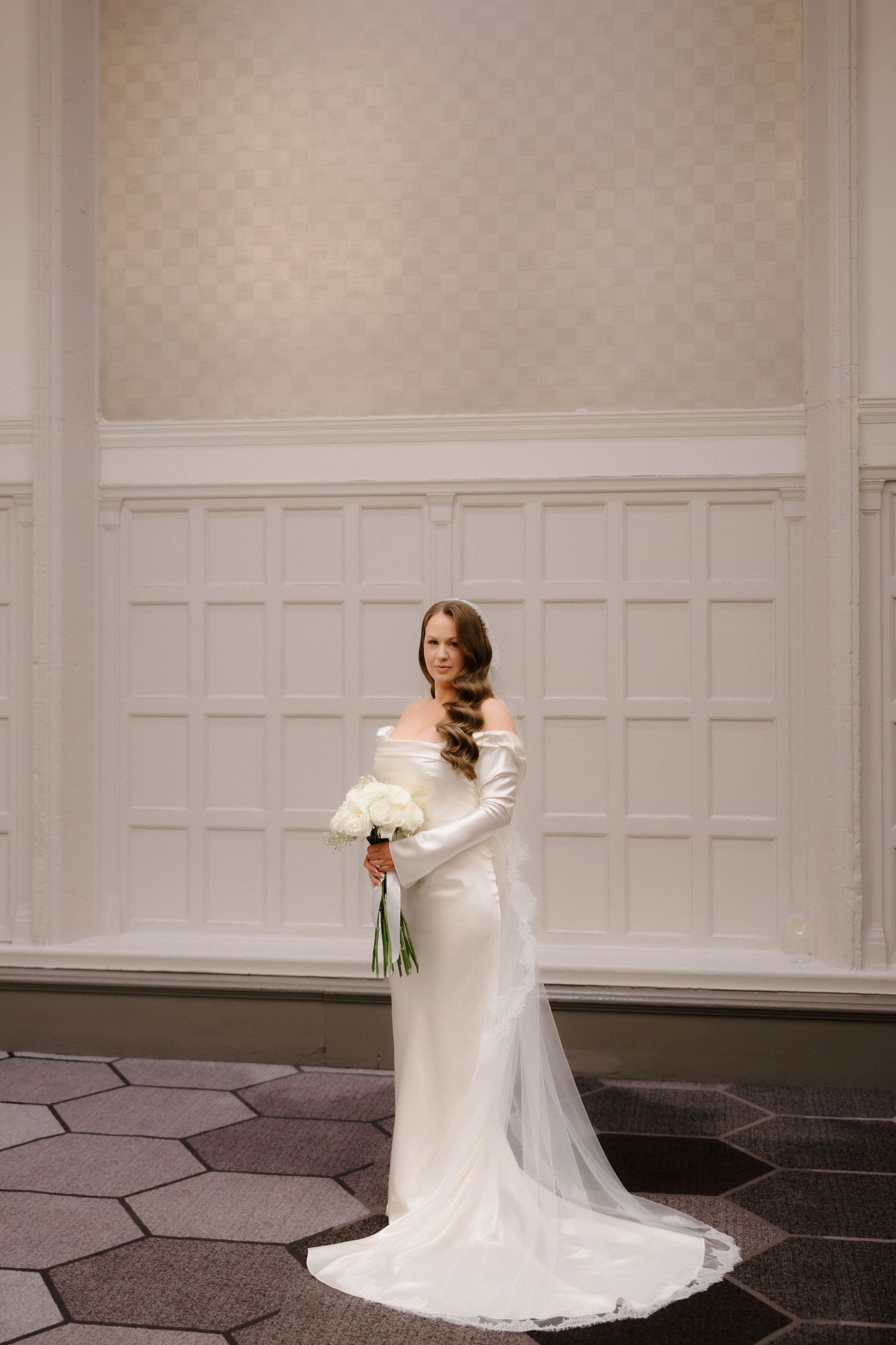 Elegant bride in a white off-shoulder gown with long train holds white bouquet in a grand ballroom.