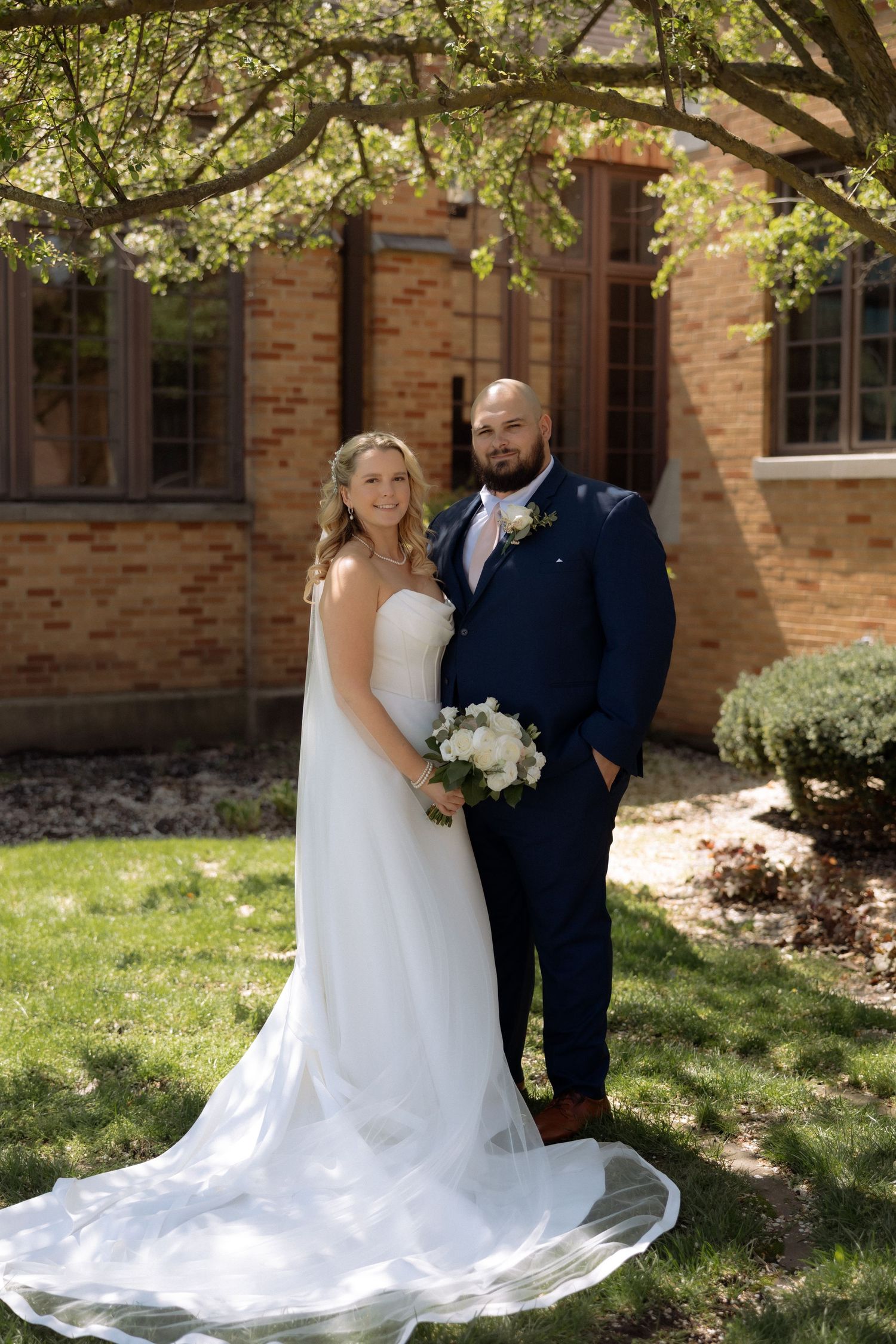 Bride in white gown and groom in navy suit pose outdoors under blooming tree near brick building on wedding day.
