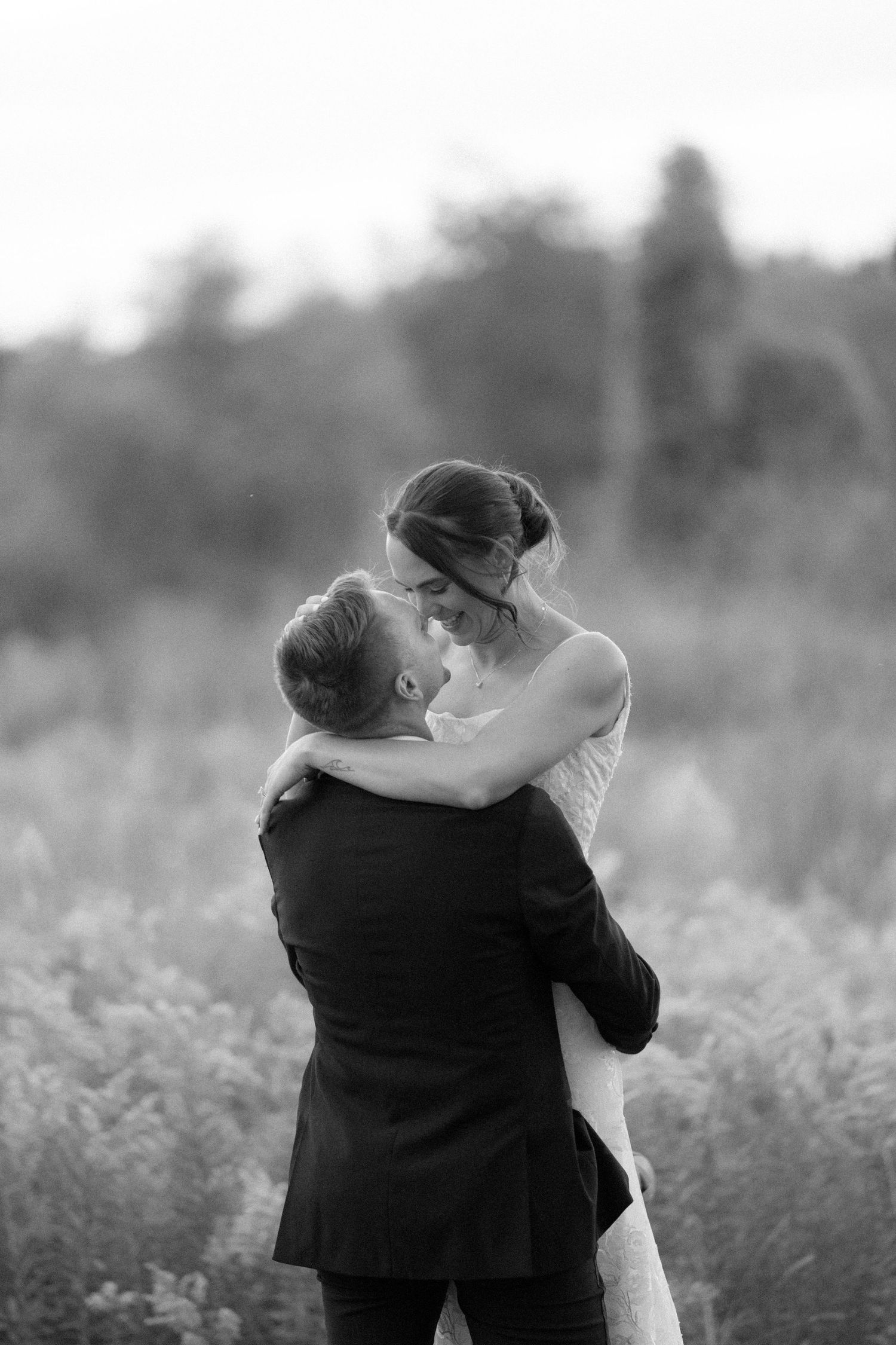 Black and white photo of groom lifting bride in an embrace amid a lush field, sharing a romantic kiss.