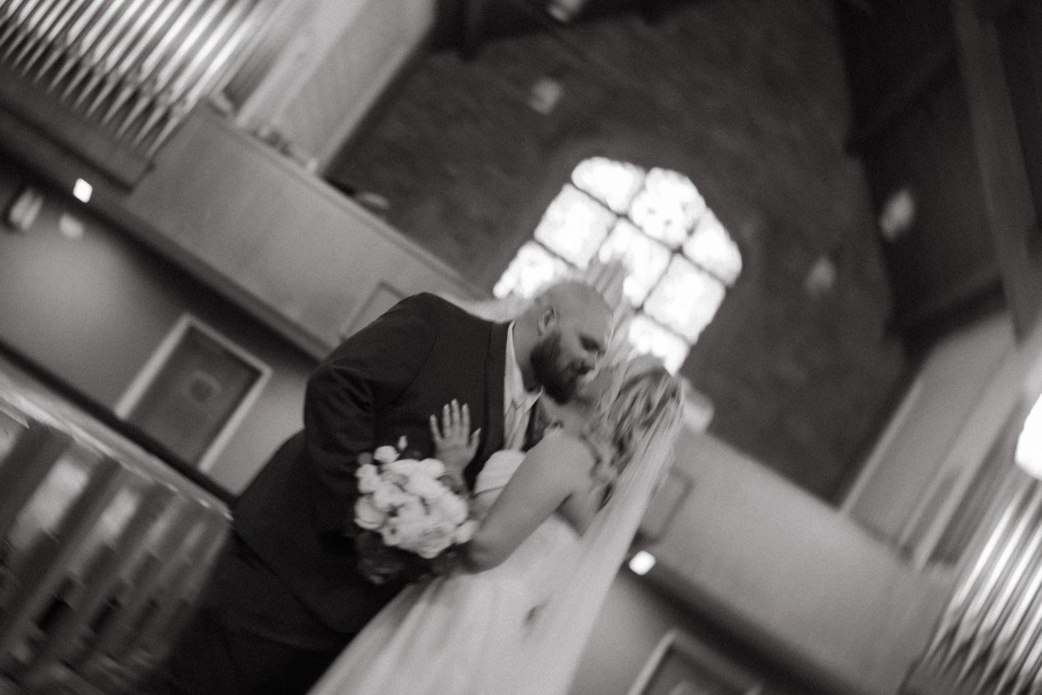 Black and white photo of a bride and groom sharing a kiss inside a church, bride holding a bouquet.