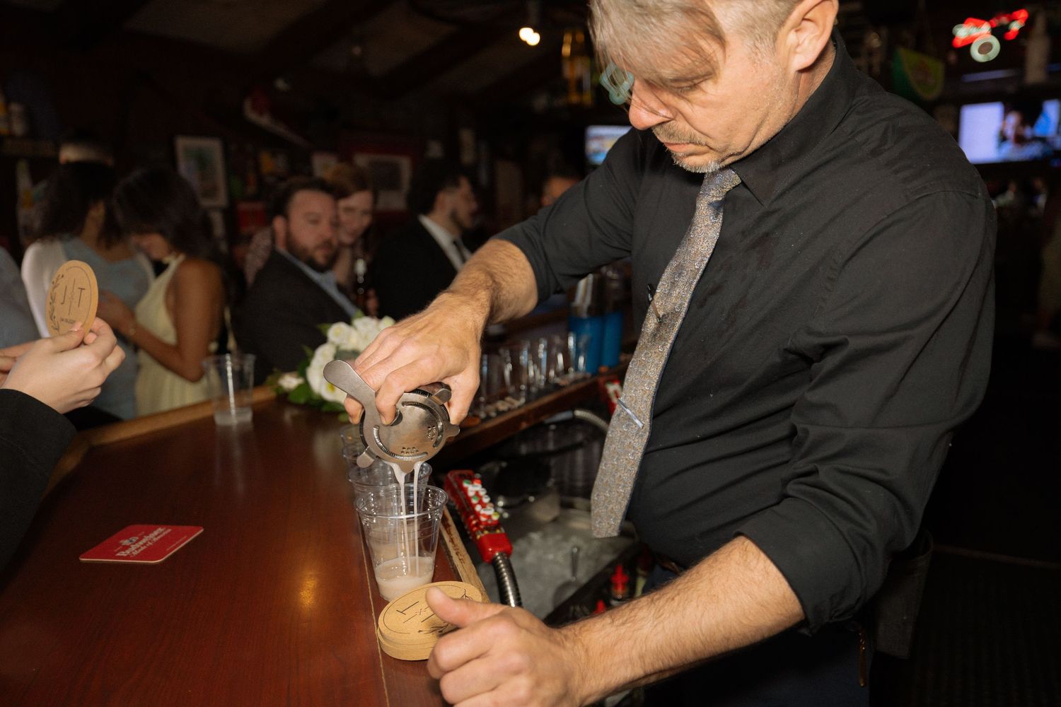 Bartender pouring a draft beer at a busy bar, wearing a black shirt and gray tie.