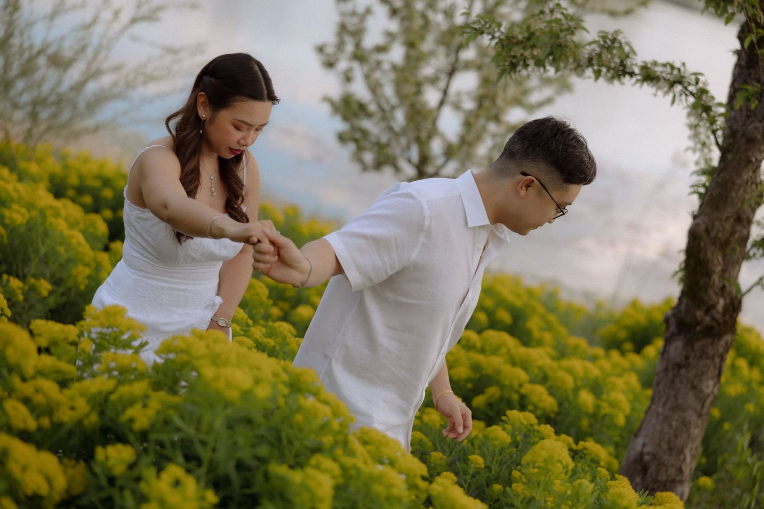 Couple in white outfits holding hands among vibrant yellow flowers in a romantic outdoor garden setting.
