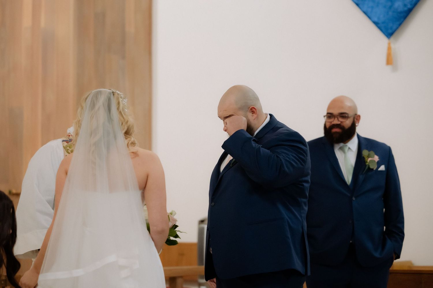 Emotional groom wiping tears as bride walks down the aisle in a white wedding dress and veil inside a church.