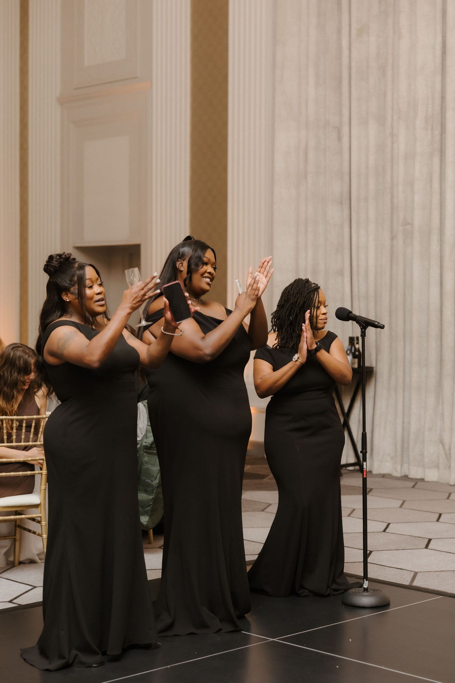 Three women in black gowns singing and clapping at a wedding reception microphone stand.