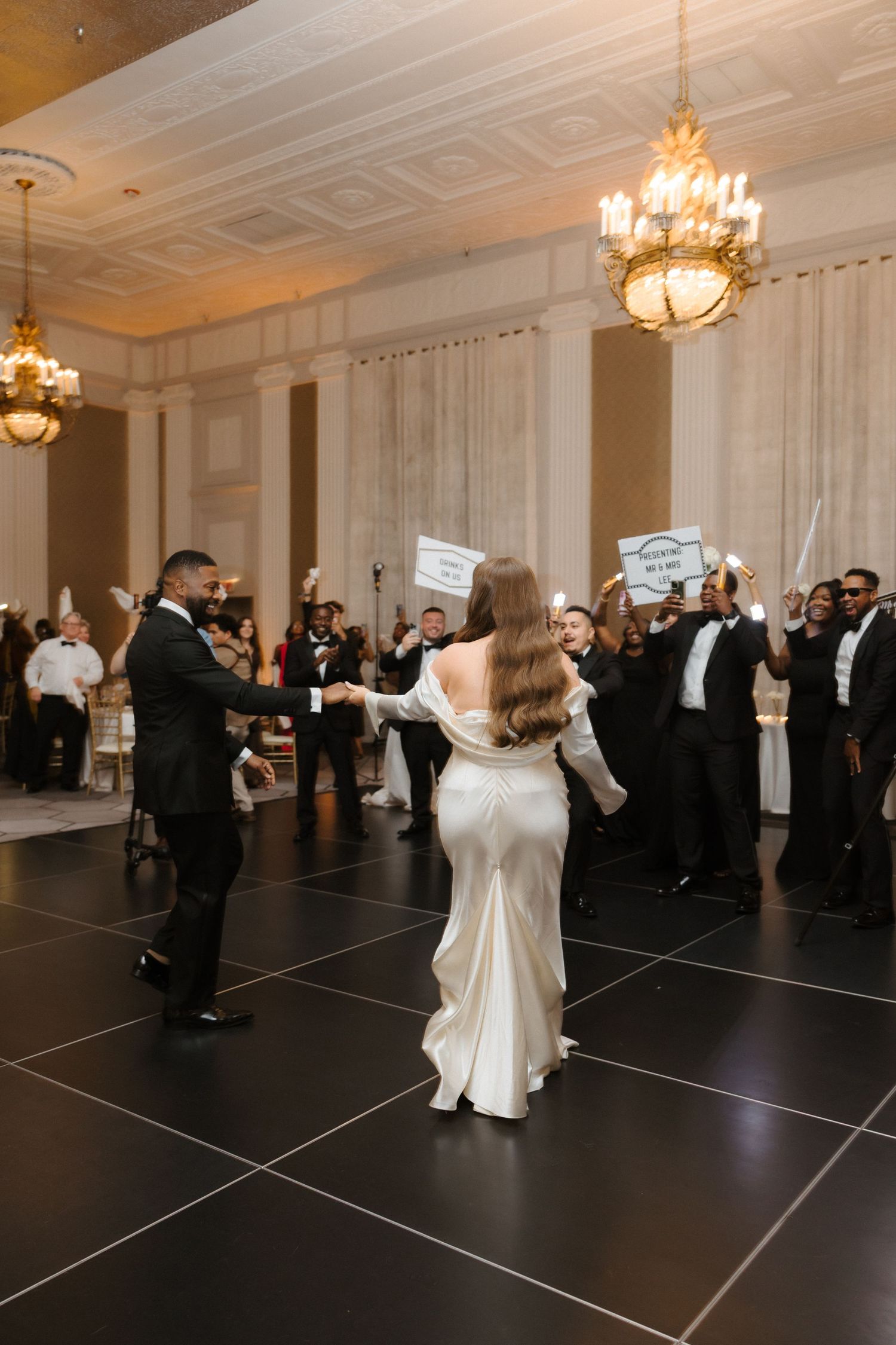 Bride and groom share first dance on black dance floor while wedding guests cheer in elegant ballroom with chandeliers.