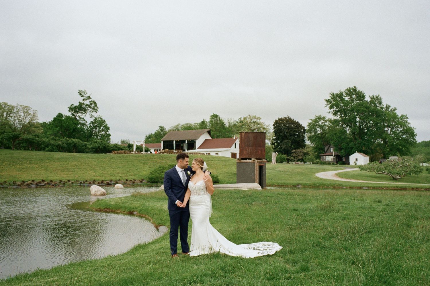 Bride and groom embrace on lush green lawn near a pond at a rustic farm wedding venue on a cloudy day.