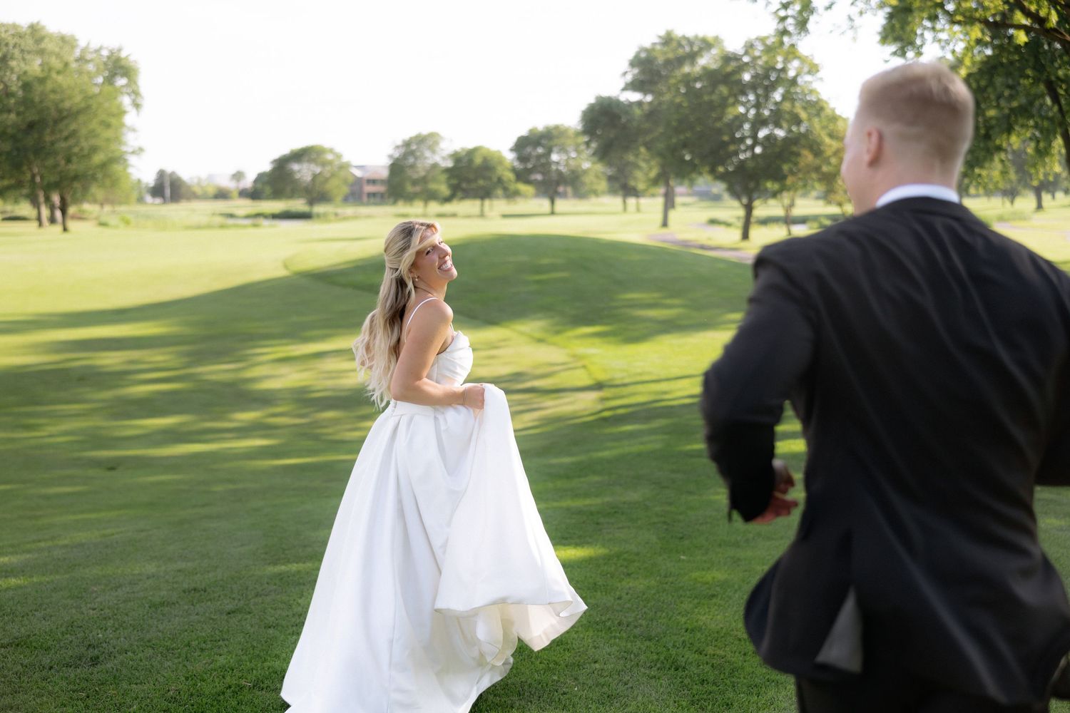 Bride in white ball gown runs toward groom across sunlit golf course green.