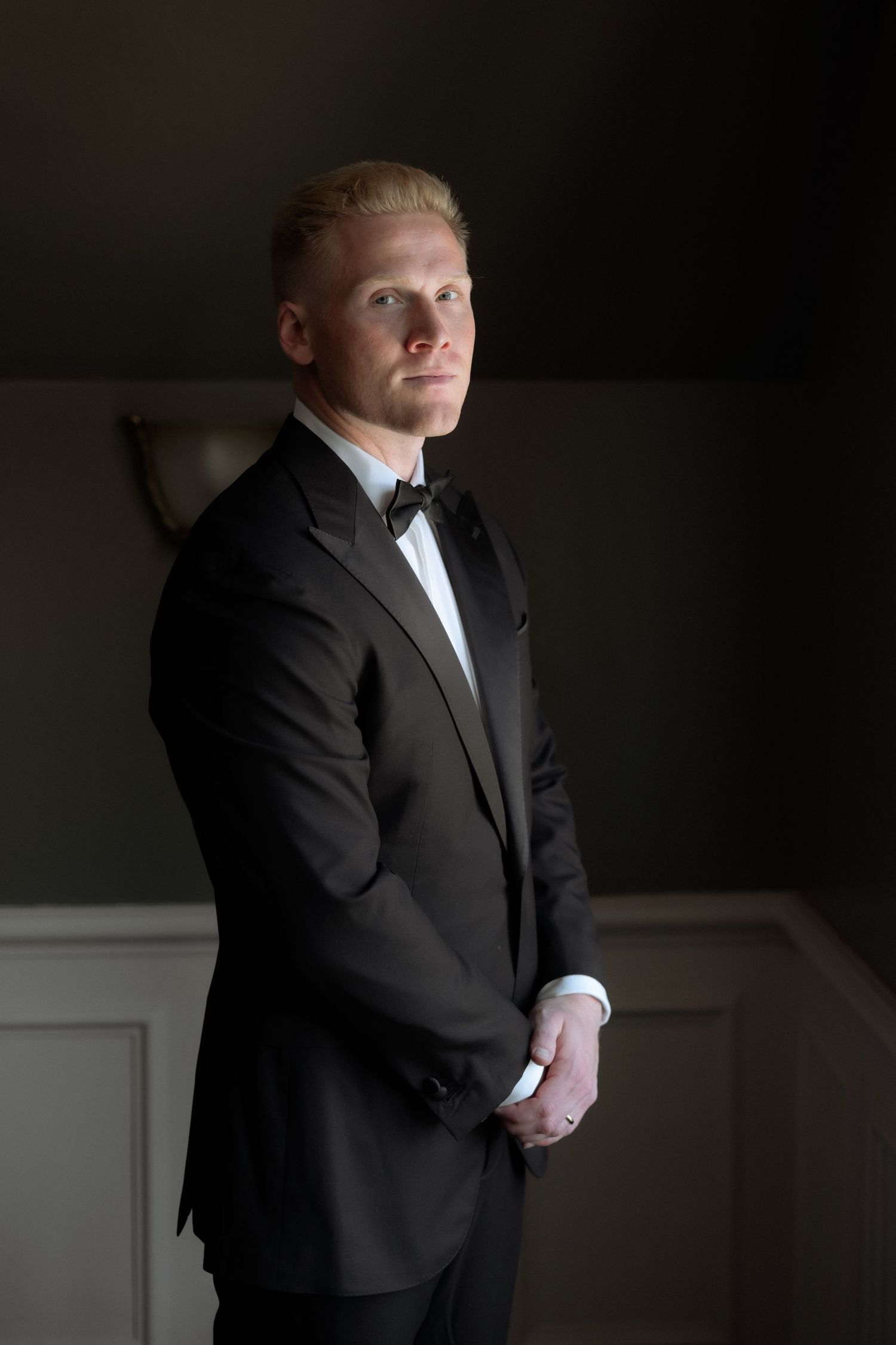 Elegant groom in black tuxedo and bow tie standing in dramatic lighting.