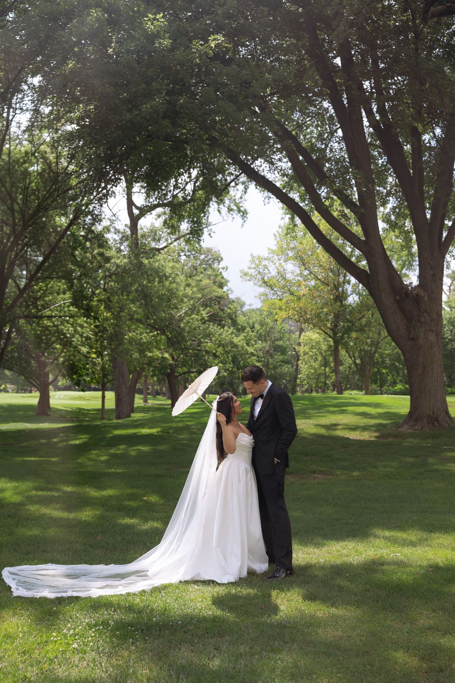 Bride and groom sharing a romantic moment under a parasol in a lush green park on their wedding day.