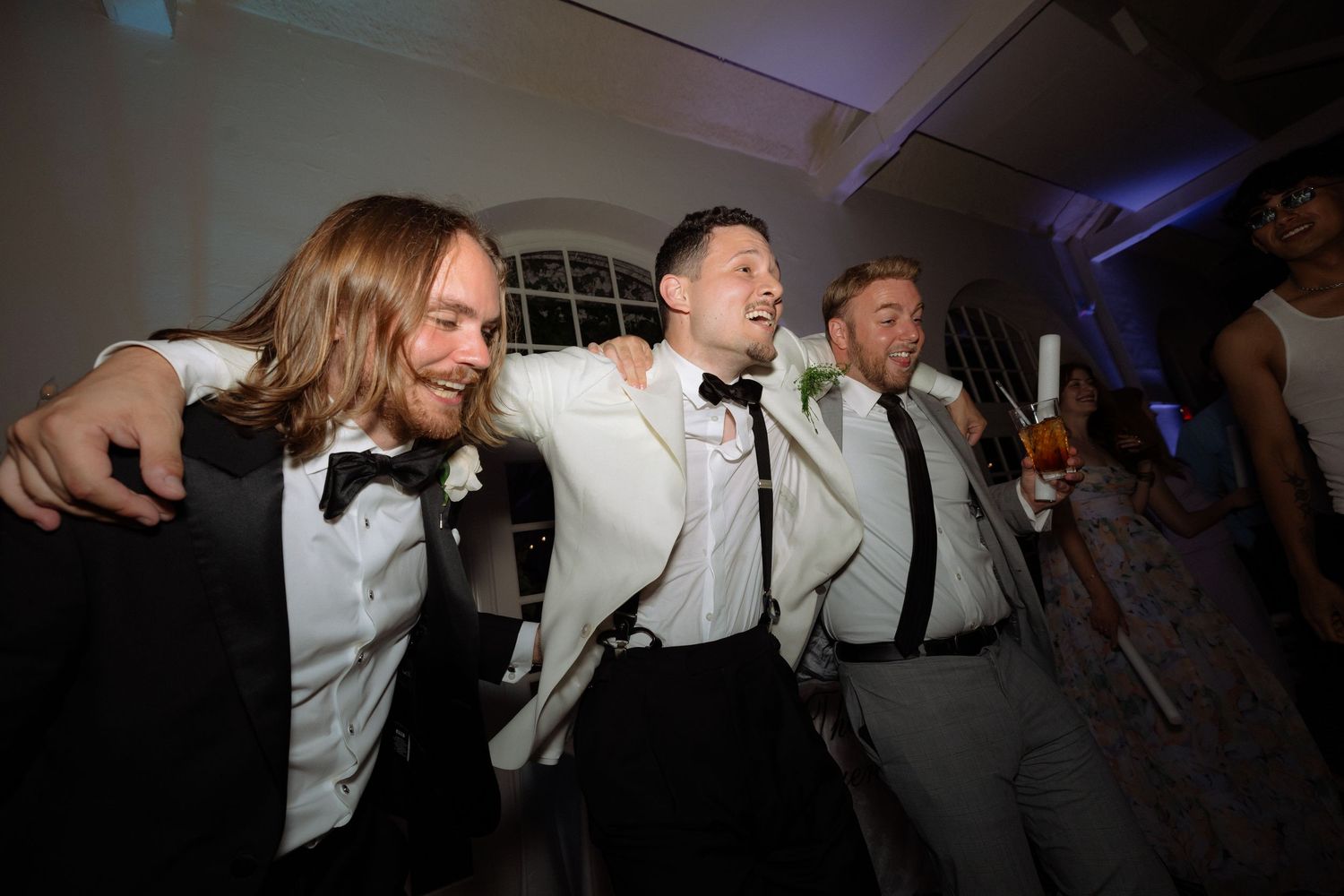 Three groomsmen in tuxedos laugh and celebrate together at wedding reception dance party.