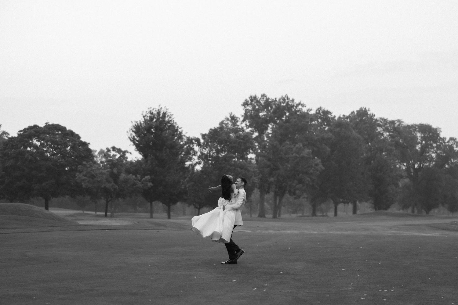 Black and white photo of groom twirling bride in white dress on open golf course.
