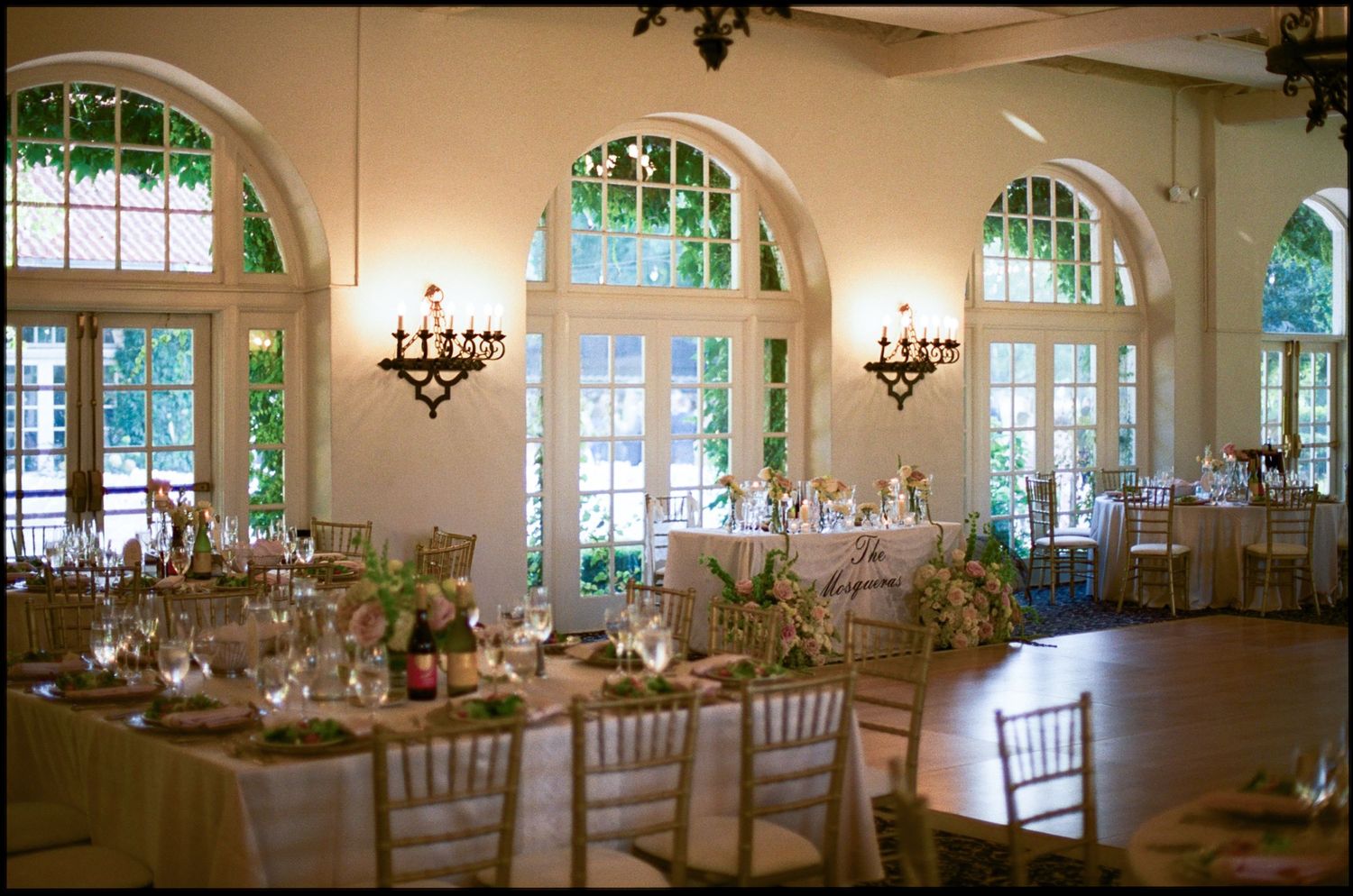Elegant wedding reception hall with arched windows, chandelier lighting, and decorated tables set for a formal dinner.