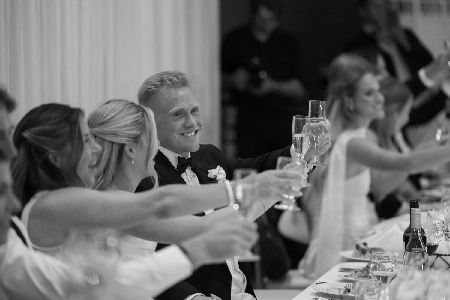 Black and white wedding toast with smiling groom and guests raising champagne glasses at elegant reception.