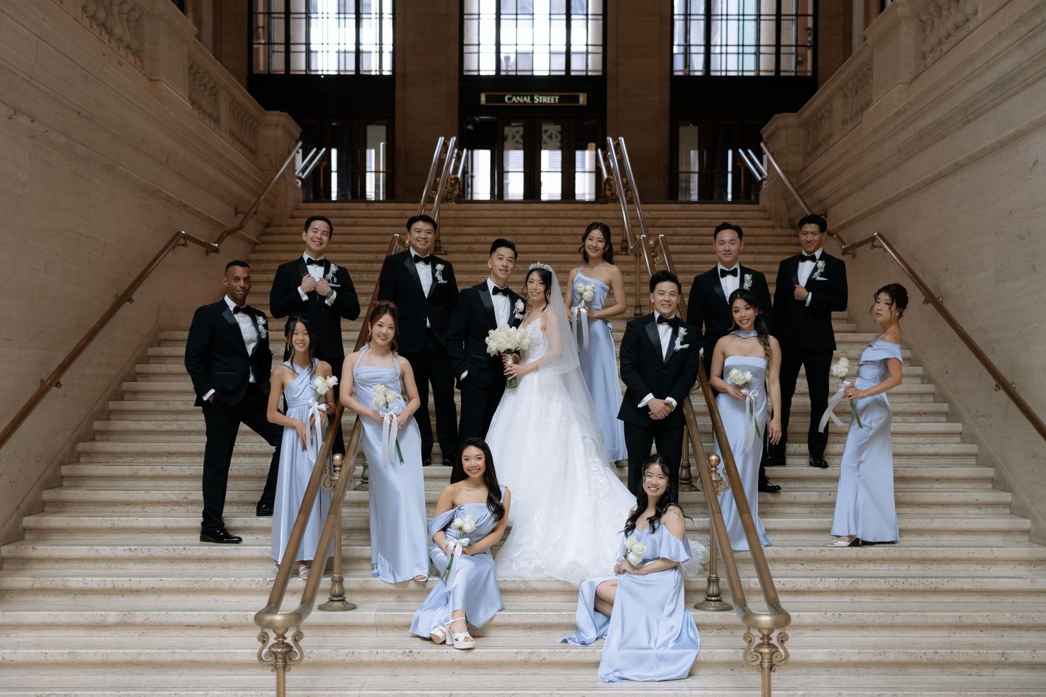 Wedding party poses on grand staircase, bride in white gown with groomsmen in tuxedos and bridesmaids in blue gowns.