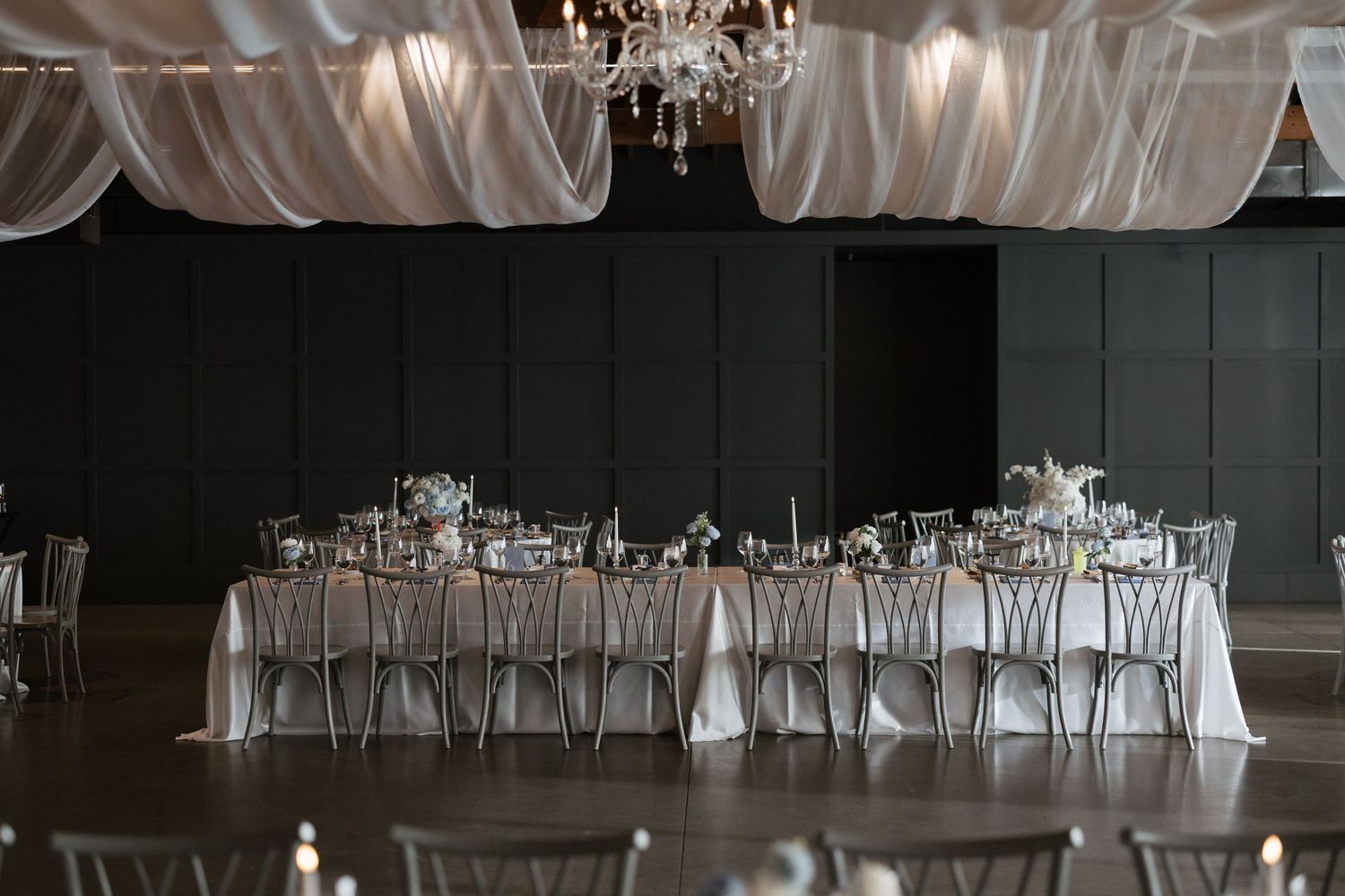 Elegant wedding reception hall with long head table, silver chairs, floral centerpieces, and draped white fabric ceiling.