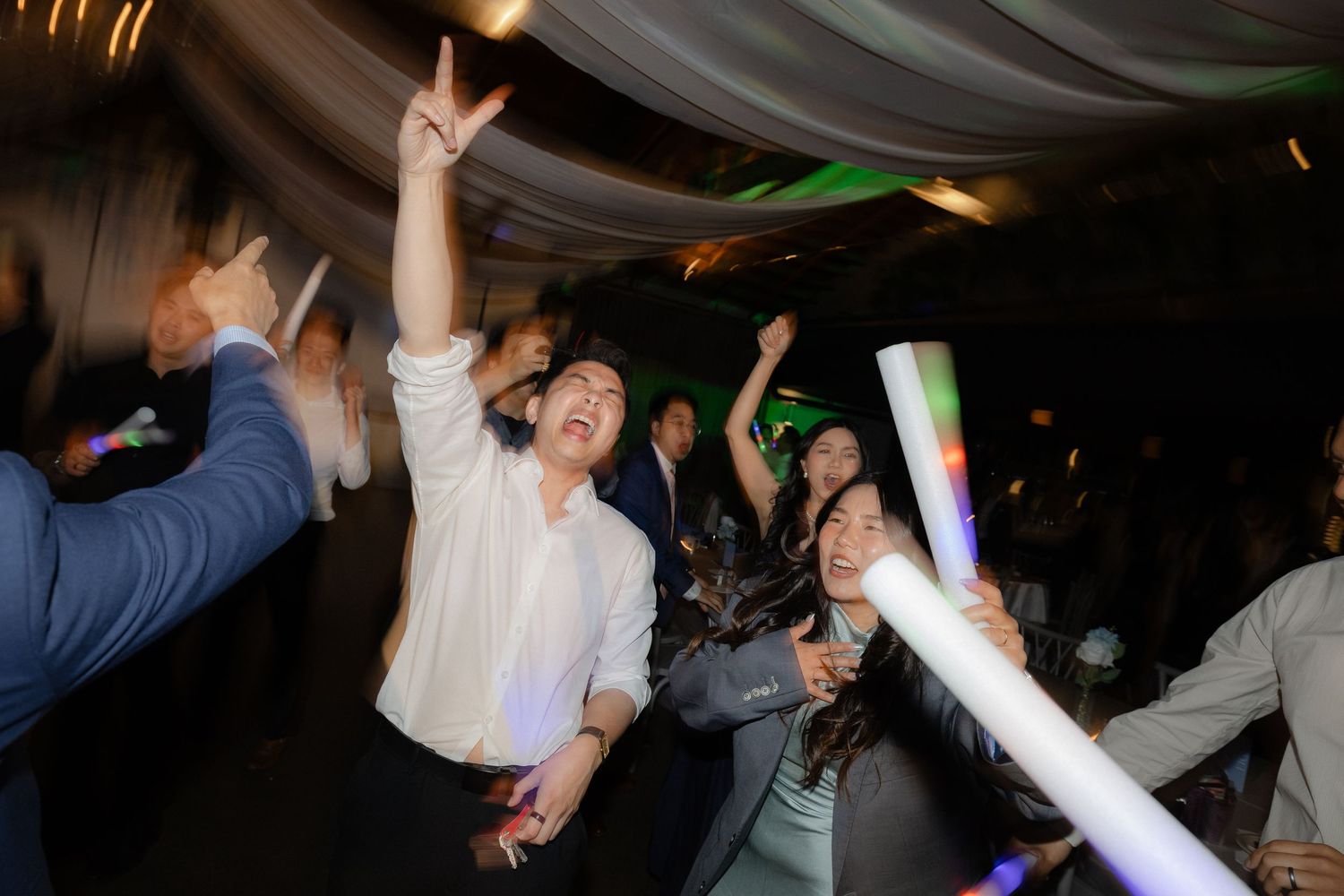 Excited wedding guests dancing and celebrating with glow sticks at energetic evening wedding reception.